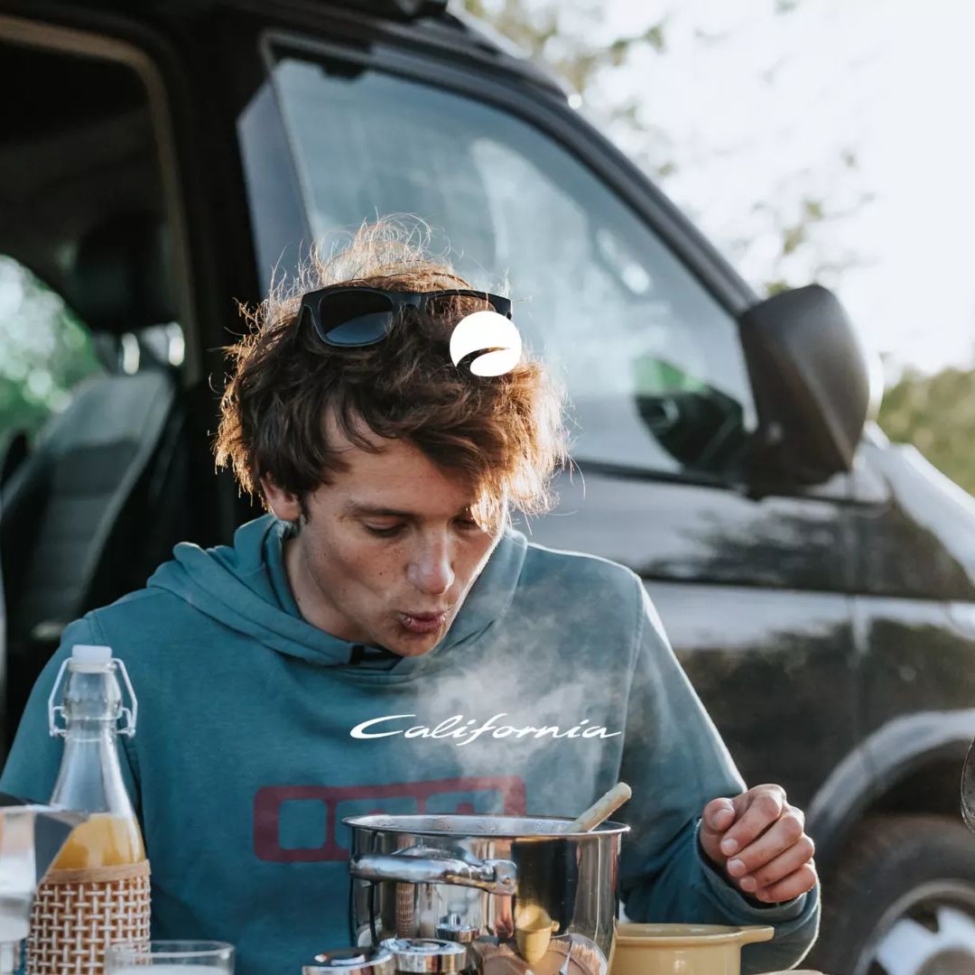 Eine Person sitzt vor einem VW California und isst an einem Tisch. Es gibt Töpfe und eine Flasche auf dem Tisch. Eine andere Person ist teilweise sichtbar im Vordergrund.