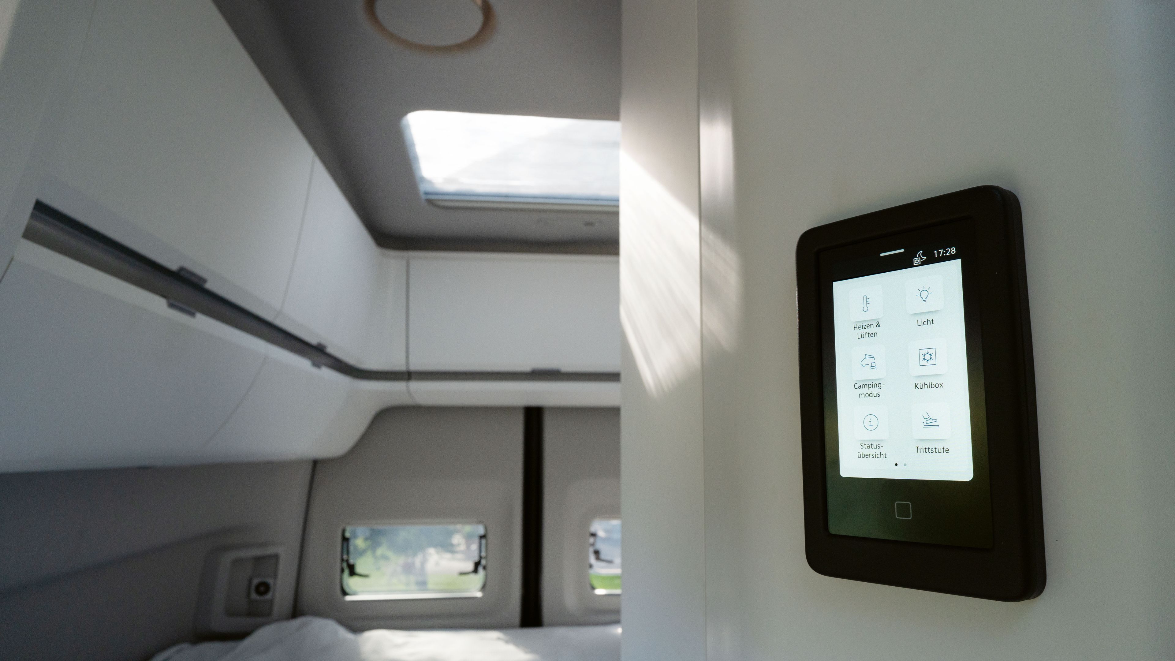 Ein modernes Wohnmobilinterieur mit einem Touchscreen-Bedienfeld an der Wand. Das Bedienfeld zeigt verschiedene Steueroptionen für Beleuchtung, Klima und andere Funktionen. Im Hintergrund sind Betten und Fenster sichtbar.
