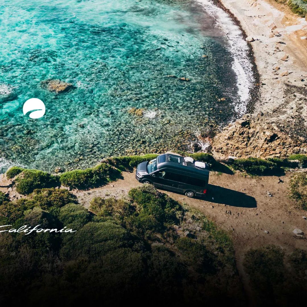 Ein grauer VW Grand California steht auf einer Klippe mit Blick auf das türkisfarbene Meer. Im Vordergrund sind Felsen und Vegetation sichtbar. Das Bild ist in Kalifornien aufgenommen.