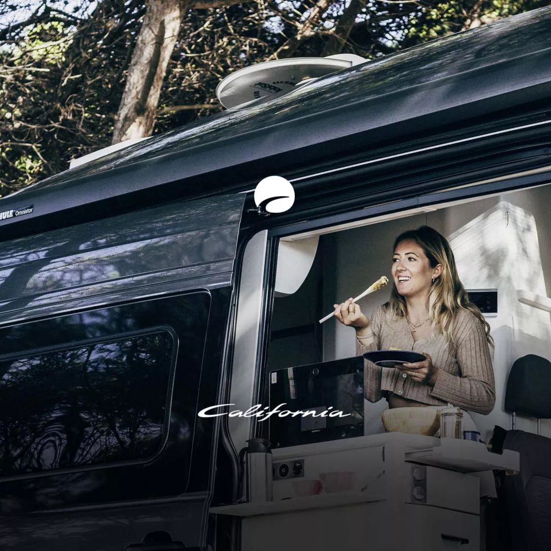 Die Frau sitzt in einem VW Grand California und isst eine Mahlzeit. Der California steht in einem Waldgebiet und hat einen modernen Aufbau. Bäume umgeben das Fahrzeug.