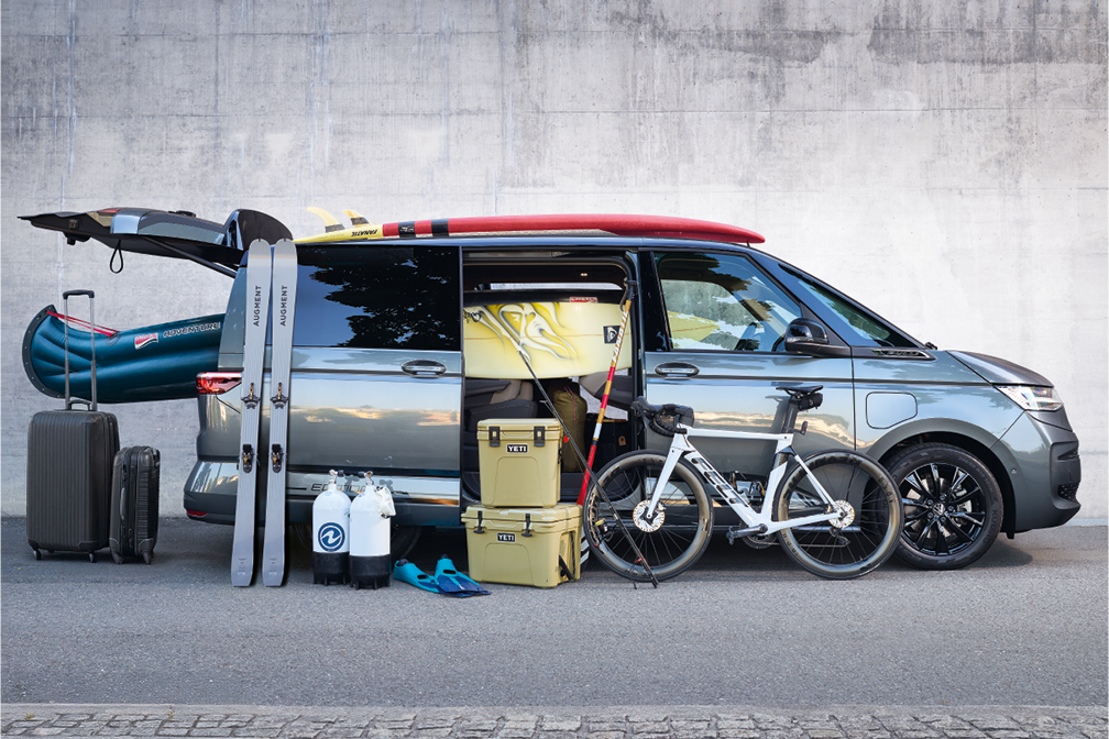 Ein grauer Van ist mit verschiedenen Freizeit- und Campingausrüstungen beladen, darunter ein Fahrrad, Tauchausrüstung, Kühlboxen, Skier, Surfbretter und ein Kanu. Der Van steht vor einer grauen Betonwand. Rechts daneben steht ein großer schwarzer Koffer.