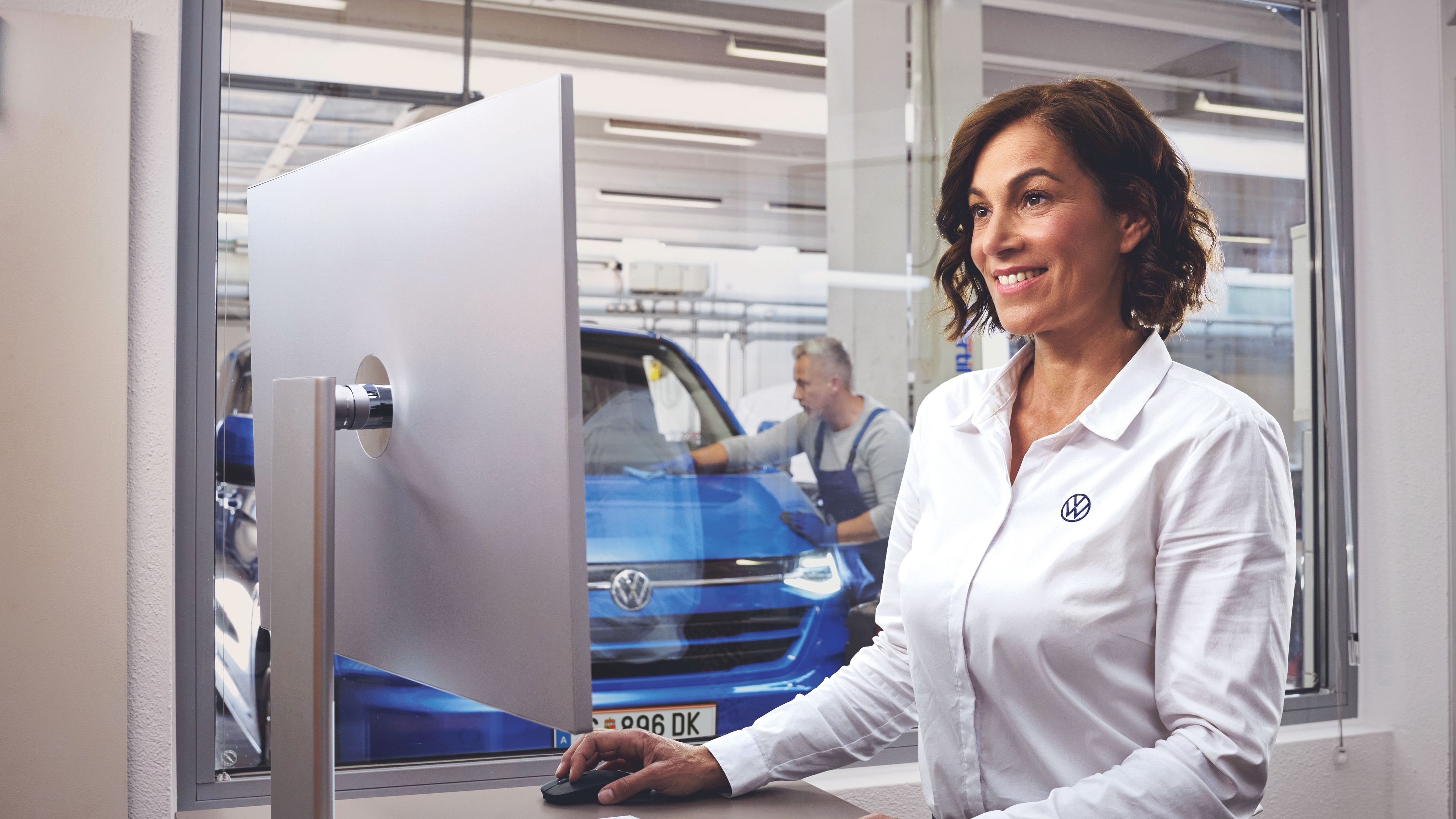 Eine Volkswagen Service MItarbeiterin tippt auf einer Tastatur und blickt auf einen Bildschirm. Im Hintergrund ist ein blauer Volkswagen Transporter in einer Werkstatt zu sehen.