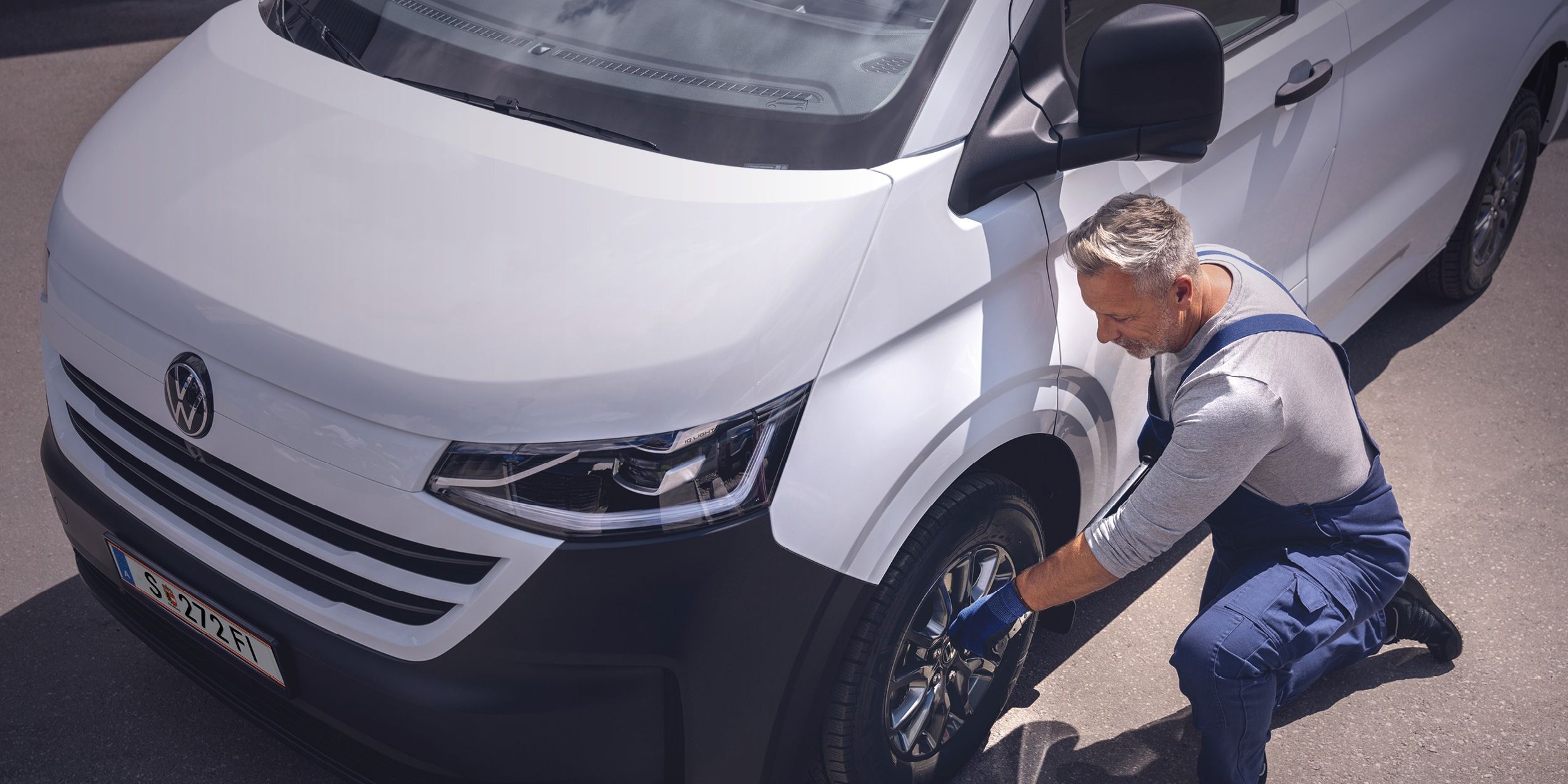 Ein Volkswagen Techniker in blauer Latzhose kniet neben einem weißen VW-Transporter und zieht mit einem Radkreuz die Radmuttern eines Vorderrads an.