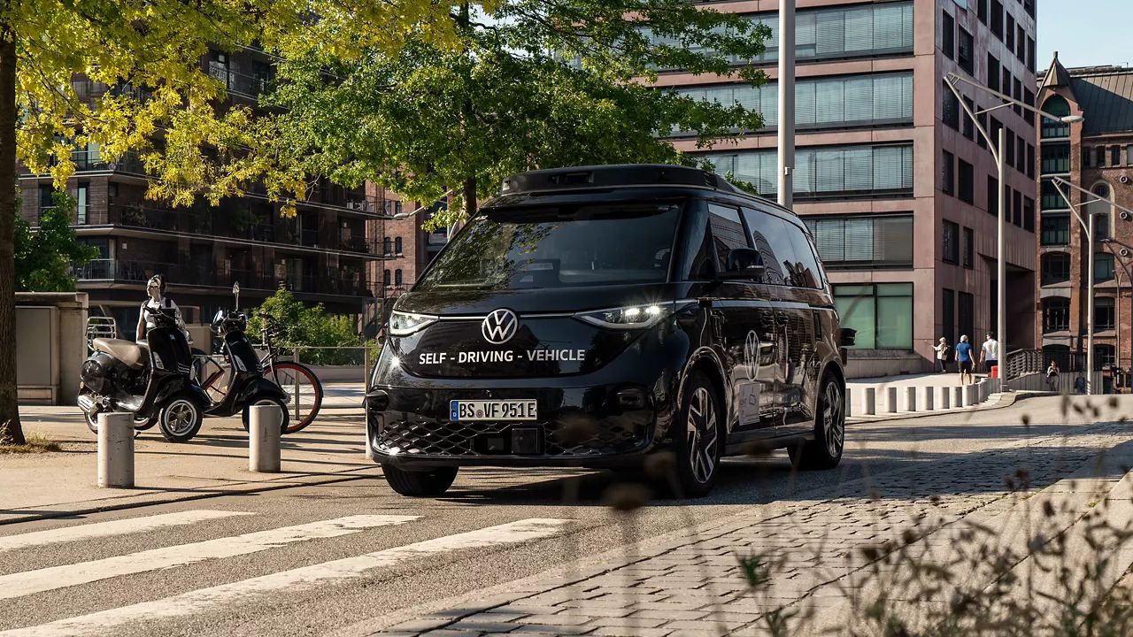 Ein schwarzes selbstfahrendes Volkswagen-Fahrzeug fährt auf einer städtischen Straße. Im Hintergrund sind moderne Gebäude und Bäume zu sehen. Neben dem Auto stehen sechs Roller nebeneinander geparkt.