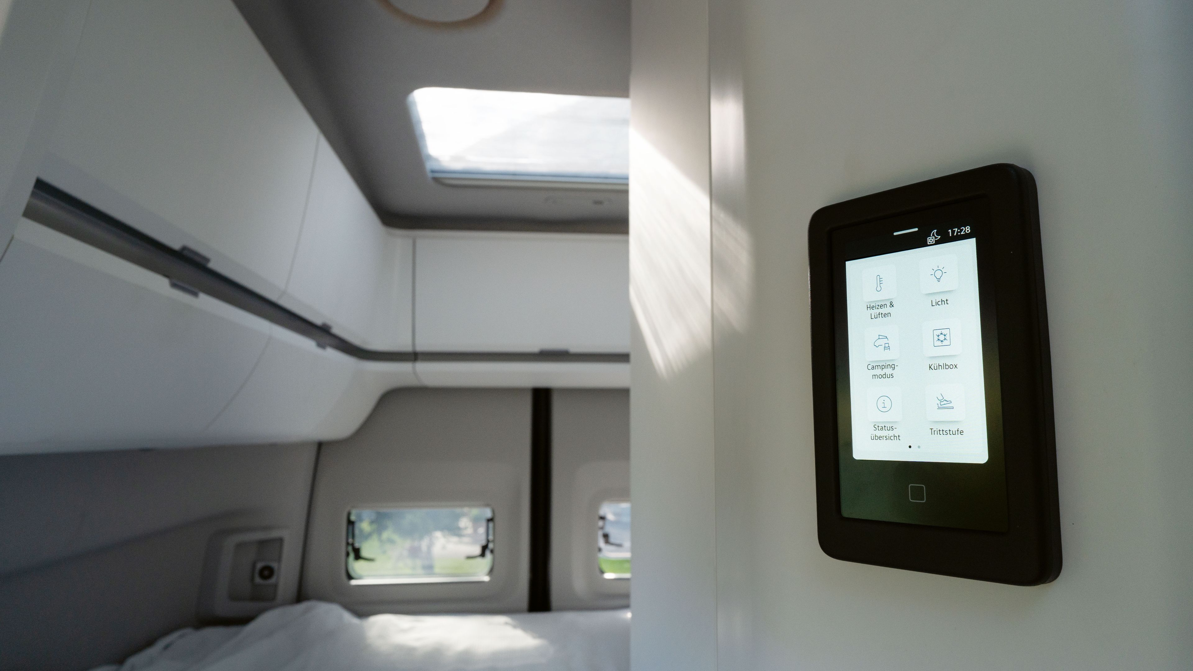 Ein modernes Wohnmobilinterieur mit einem Touchscreen-Bedienfeld an der Wand. Das Bedienfeld zeigt verschiedene Steueroptionen für Beleuchtung, Klima und andere Funktionen. Im Hintergrund sind Betten und Fenster sichtbar.