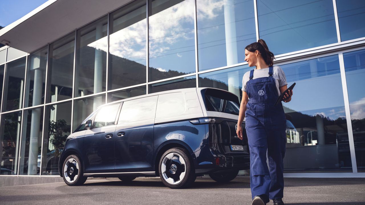 Ein blauer ID. Buzz Cargo steht vor einer Volkswagen Werkstatt. Eine Volkswagen Technikerin mit Tablet in der Hand bewegt sich von dem Fahrzeug fort.