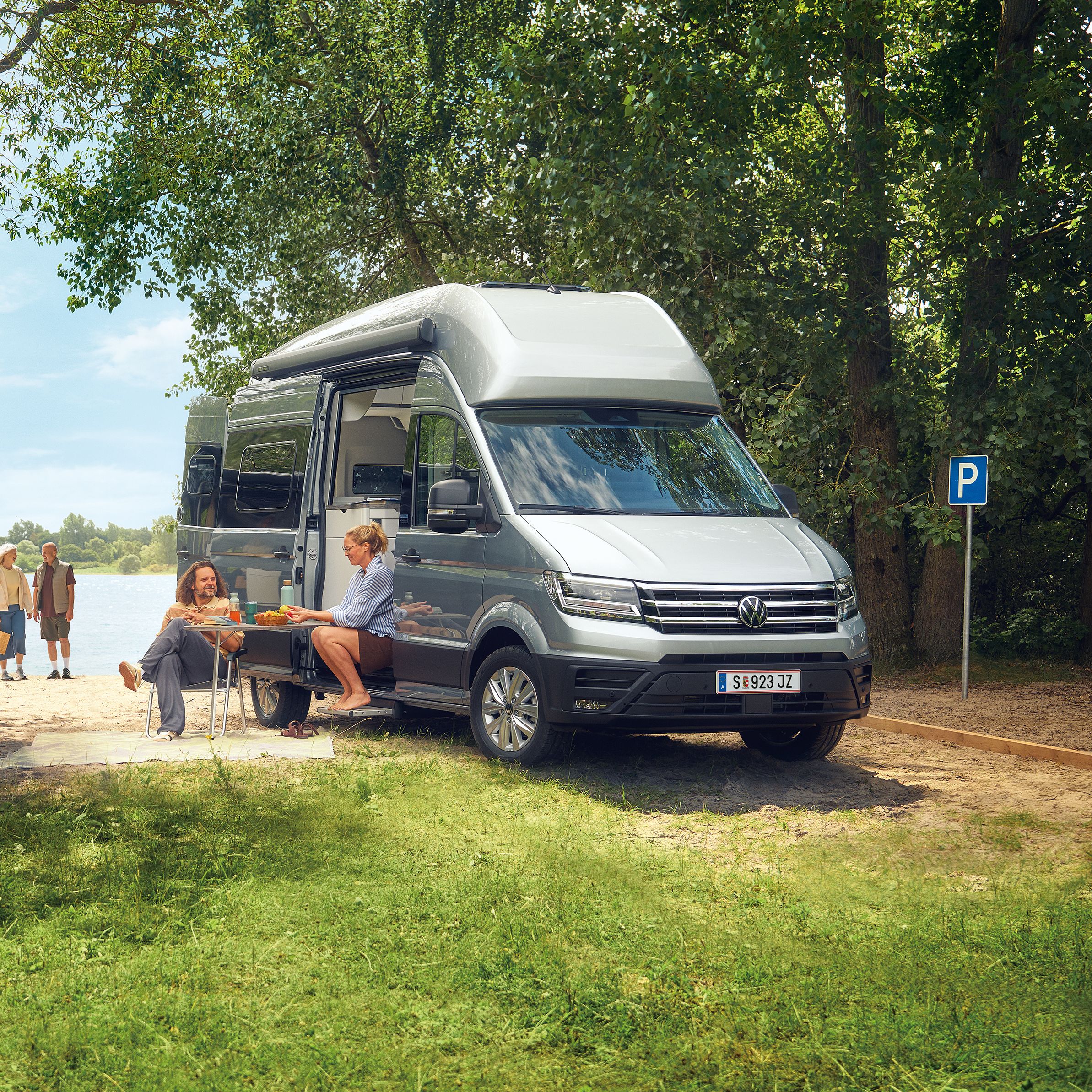 Zwei VW Grand California stehen auf einer grasbewachsenen Fläche in der Nähe eines Sees. Es gibt mehrere Personen; eine sitzt an einem Picknicktisch und zwei weitere gehen auf das Wasser zu. Die Szene ist von Bäumen und Grün umgeben.