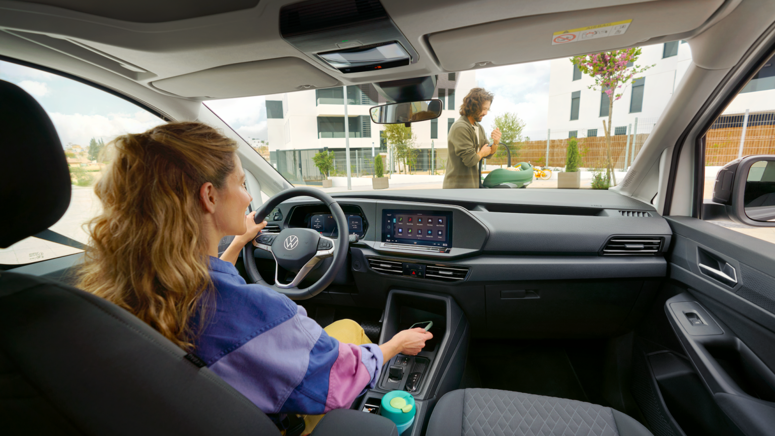 Innenraum eines VW Caddy, Frau sitzt am Steuer und legt ihr Handy in die Mittelkonsole, Mann mit Baby geht vor dem Auto