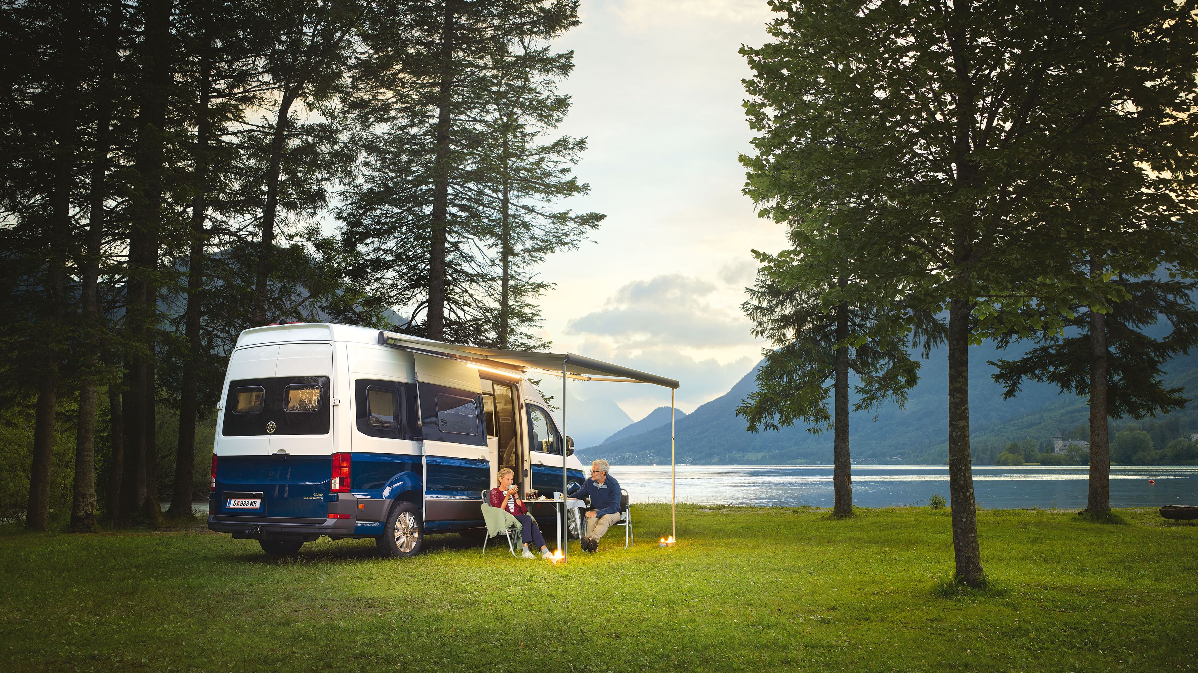 Der Grand California in der Abenddämmerung vor einem See. Ein älteres Paar sitzt unter der Markise und trinkt eine Tasse Tee. 