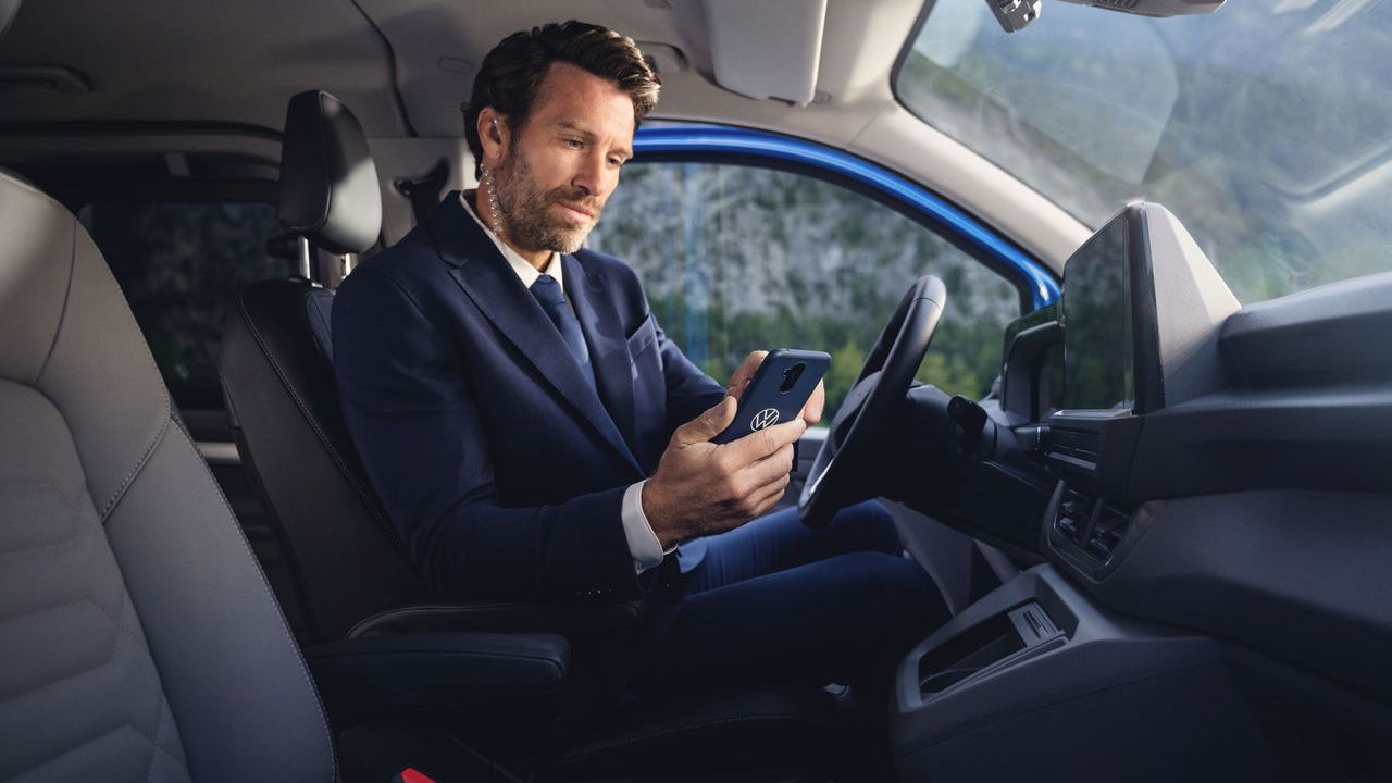 Ein Mann im Anzug sitzt auf dem Fahrersitz eines Volkswagen Nutzfahrzeuges und blickt konzentriert auf sein Smartphone in einer Hülle mit Volkswagen Logo.