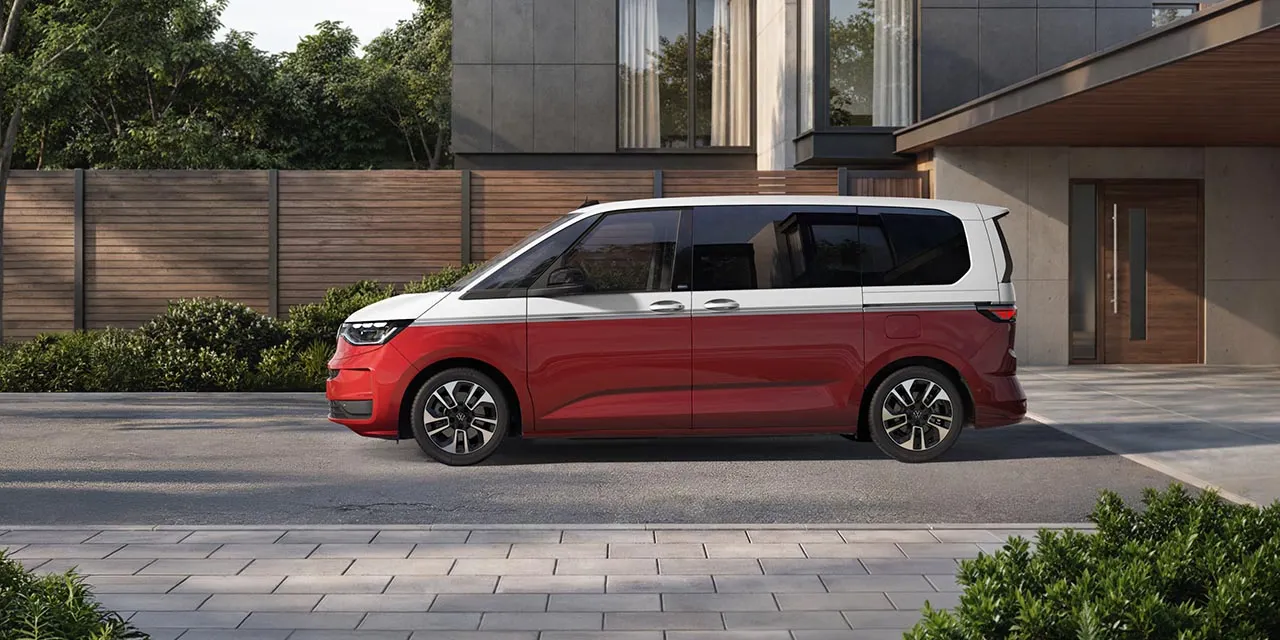 Ein rot weißer Volkswagen Multivan steht seitlich auf einer gepflasterten Einfahrt vor einem schicken, minimalistischen Wohnhaus.