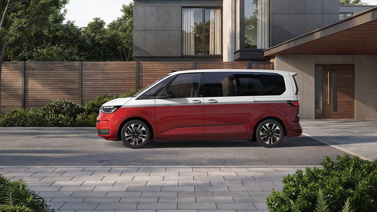 Ein rot weißer Volkswagen Multivan steht seitlich auf einer gepflasterten Einfahrt vor einem schicken, minimalistischen Wohnhaus.