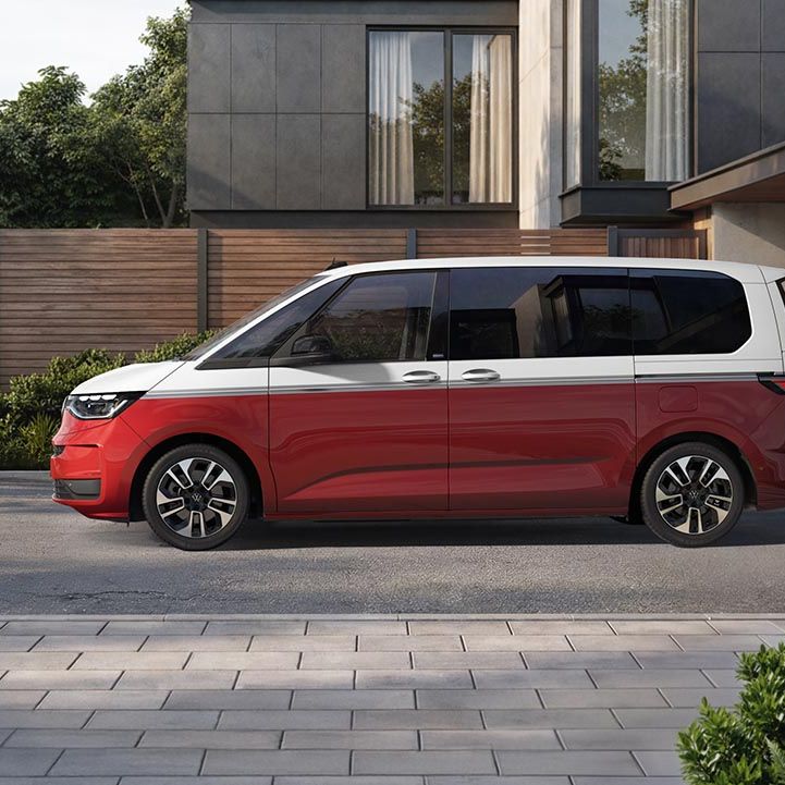 Ein rot weißer Volkswagen Multivan steht seitlich auf einer gepflasterten Einfahrt vor einem schicken, minimalistischen Wohnhaus.