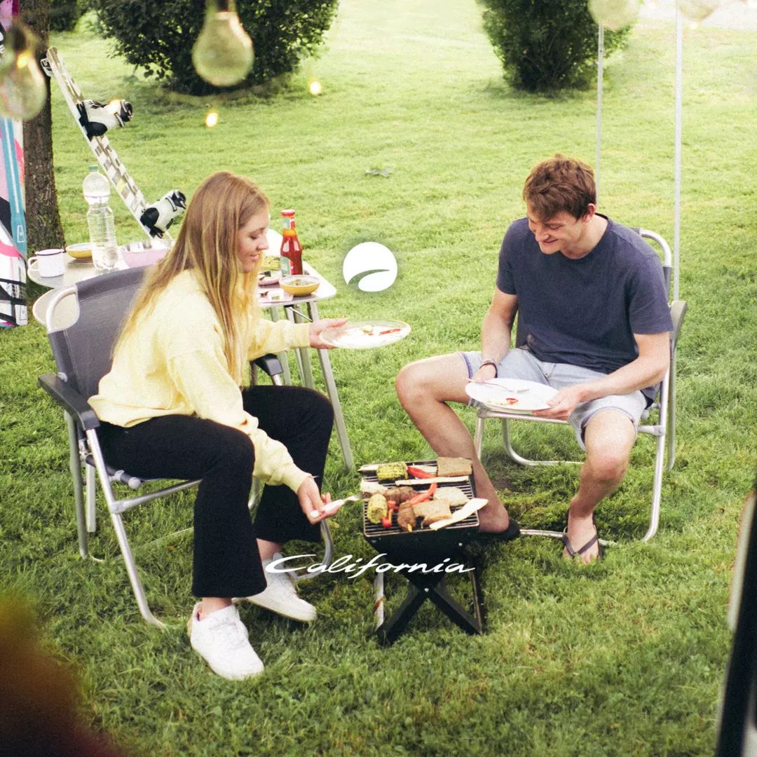 Eine Frau und ein Mann sitzen auf Campingstühlen im Freien. Sie haben einen kleinen Tisch mit Lebensmitteln zwischen sich. Im Hintergrund stehen ein buntes Zelt und ein Kleinbus.