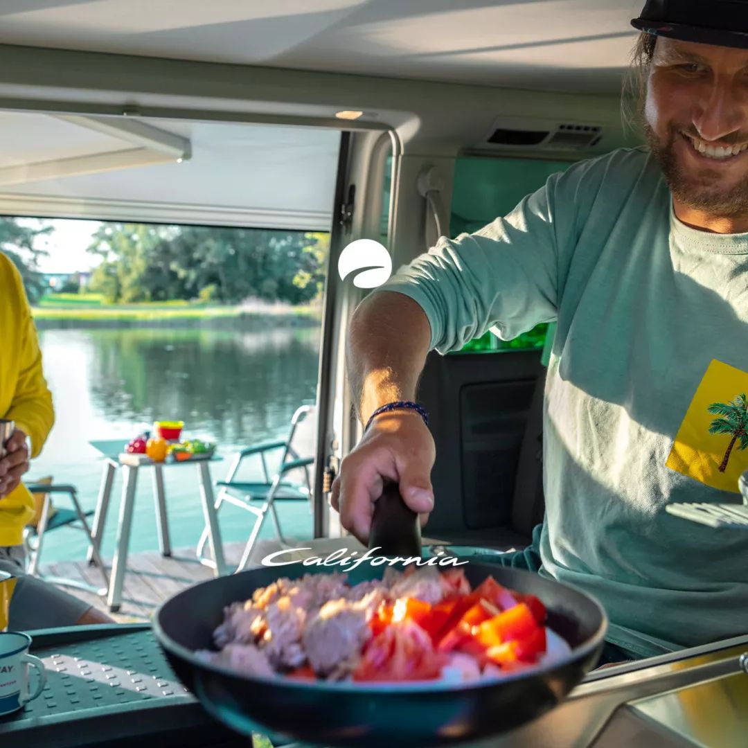 Zwei Männer sitzen in einem Wohnmobil und genießen eine Mahlzeit, die auf einem kochenden Herd zubereitet wird. Im Hintergrund sieht man einen See und grünes Gras. Auf dem Tisch draußen stehen Getränke und Snacks.