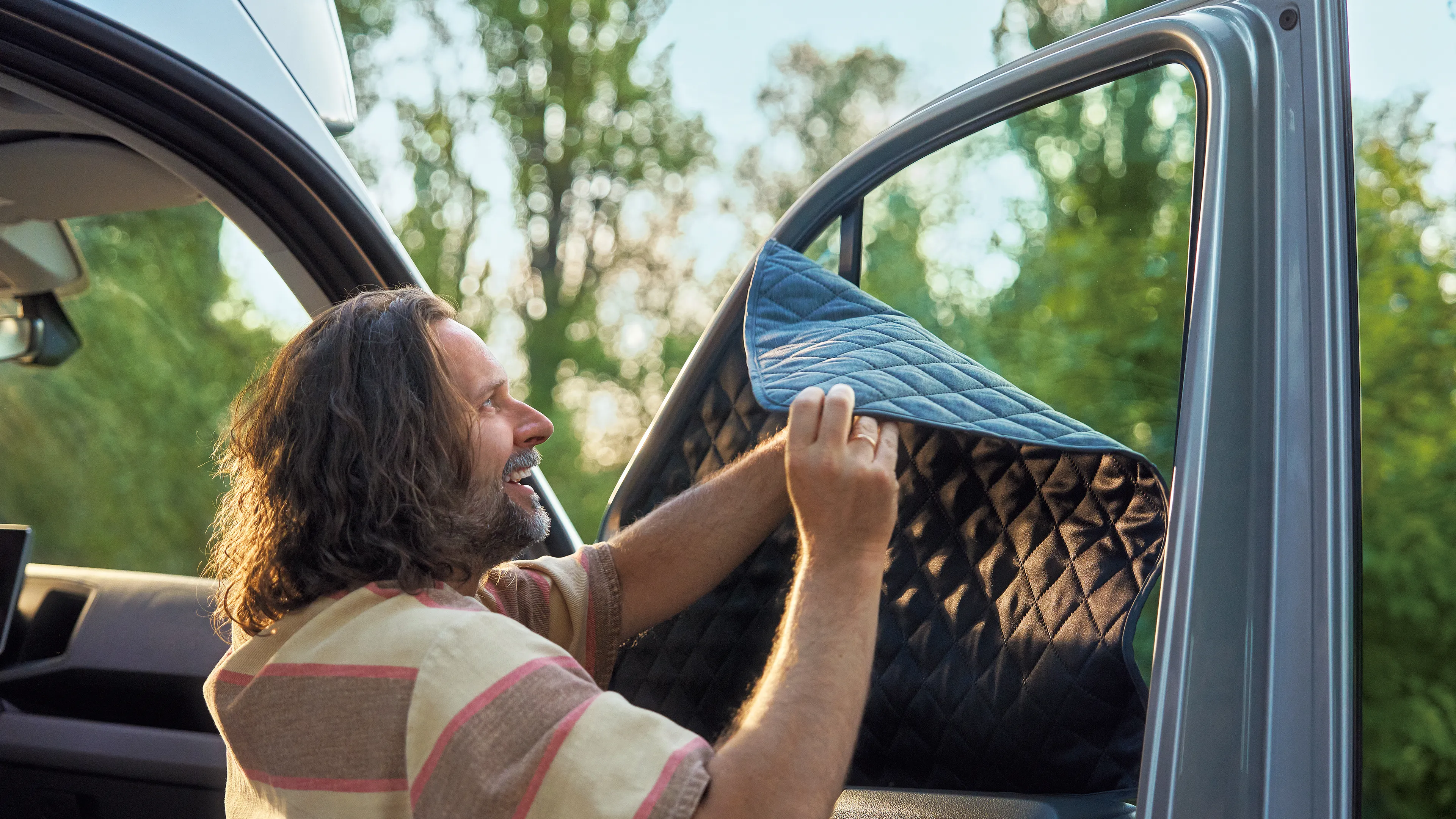 Eine Person mit langen Haaren befestigt eine isolierende Decke an der Innenseite der Fahrertür eines Vans. Die Umgebung deutet auf einen Camping- oder Outdoor-Umfeld hin.