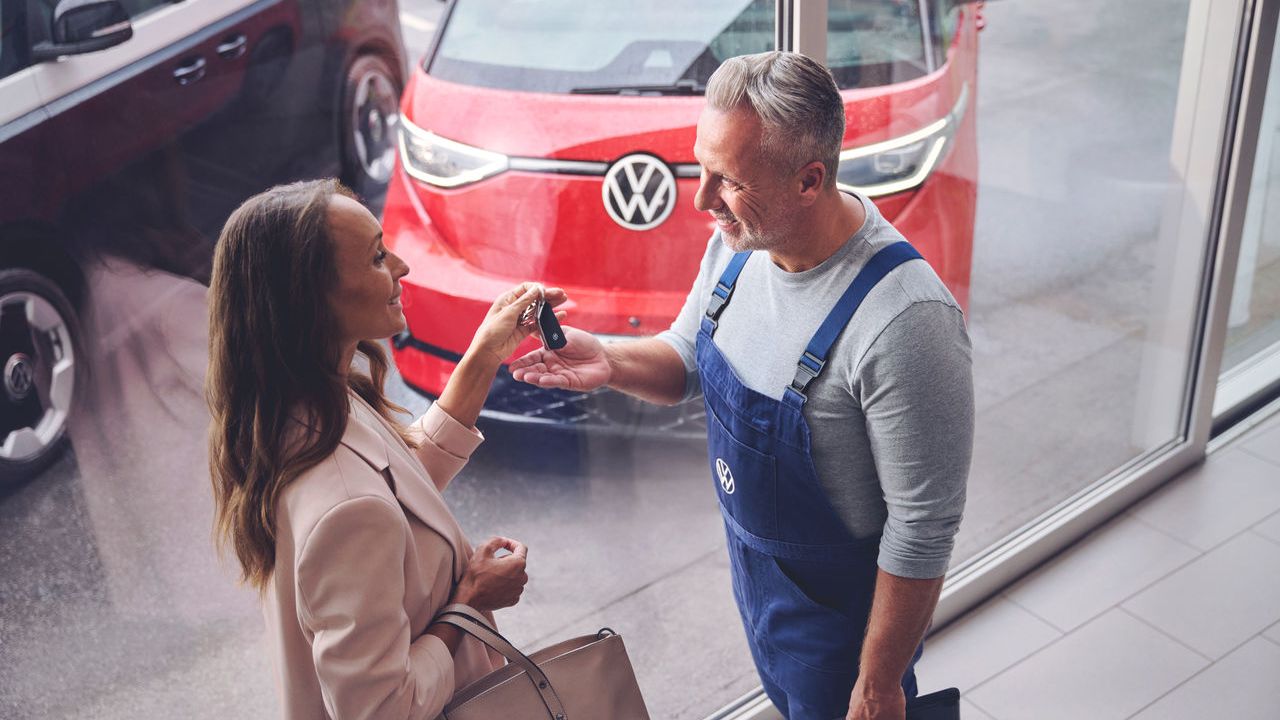 Ein Volkswagen Techniker überreicht einer Kundin ihren Autoschlüssel. Im Hintergrund befindet sich ein roter Volkswagen ID. Buzz.