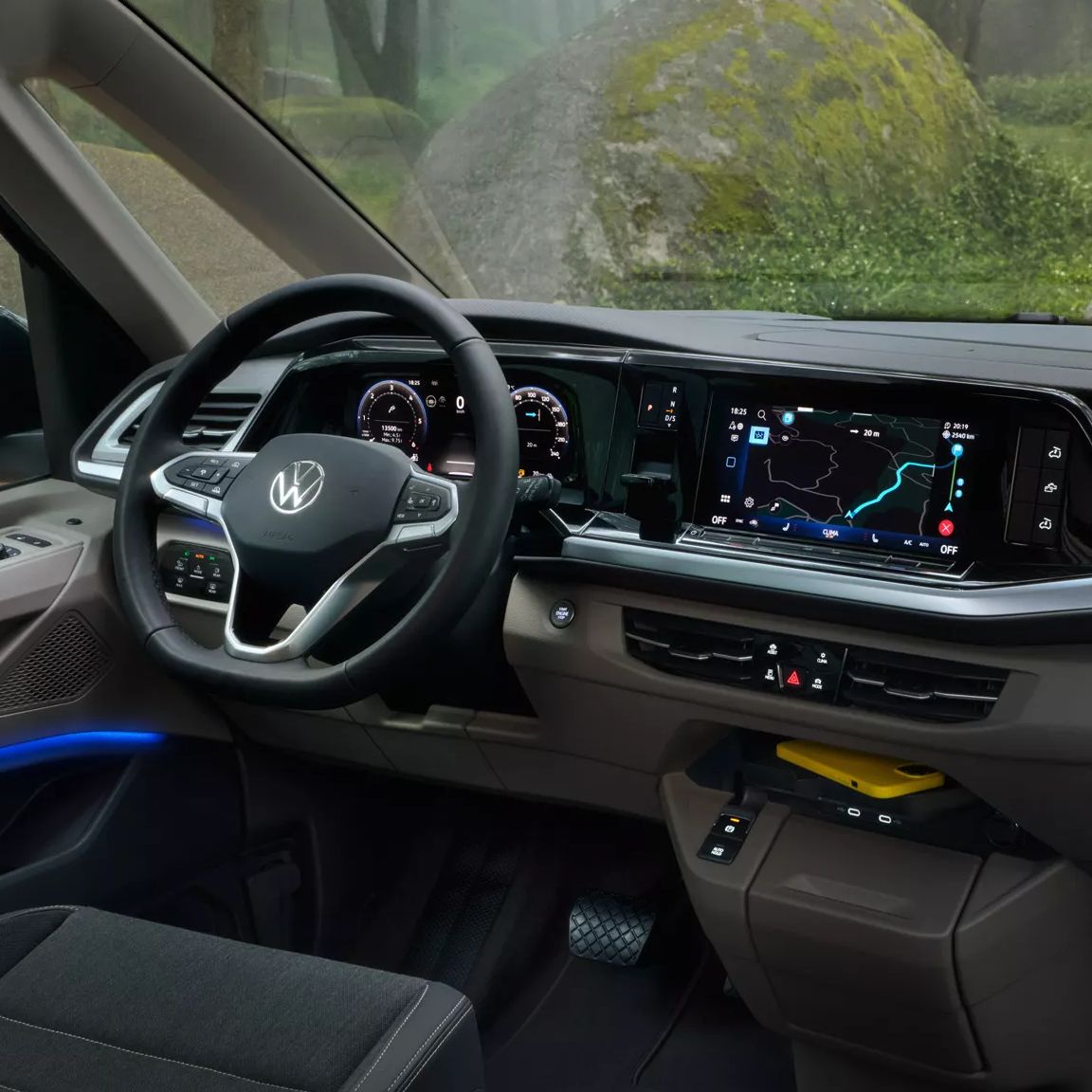 Das Bild zeigt das Innere eines Grand California, dessen Mittelpunkt der moderne Armaturenbrettbereich ist. Im Hintergrund sind zwei Personen mit Rucksäcken zu sehen, die in einem waldähnlichen Gebiet spazieren gehen. Moderne Technik wie Navigationssysteme und LED-Beleuchtung sind klar erkennbar.