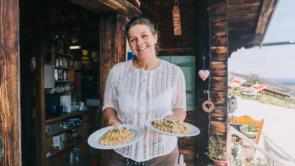 Eine Frau steht vor einer rustikalen Berghütte und hält zwei Teller mit Essen in den Händen. Im Hintergrund sind Holzmöbel und eine Berglandschaft sichtbar.