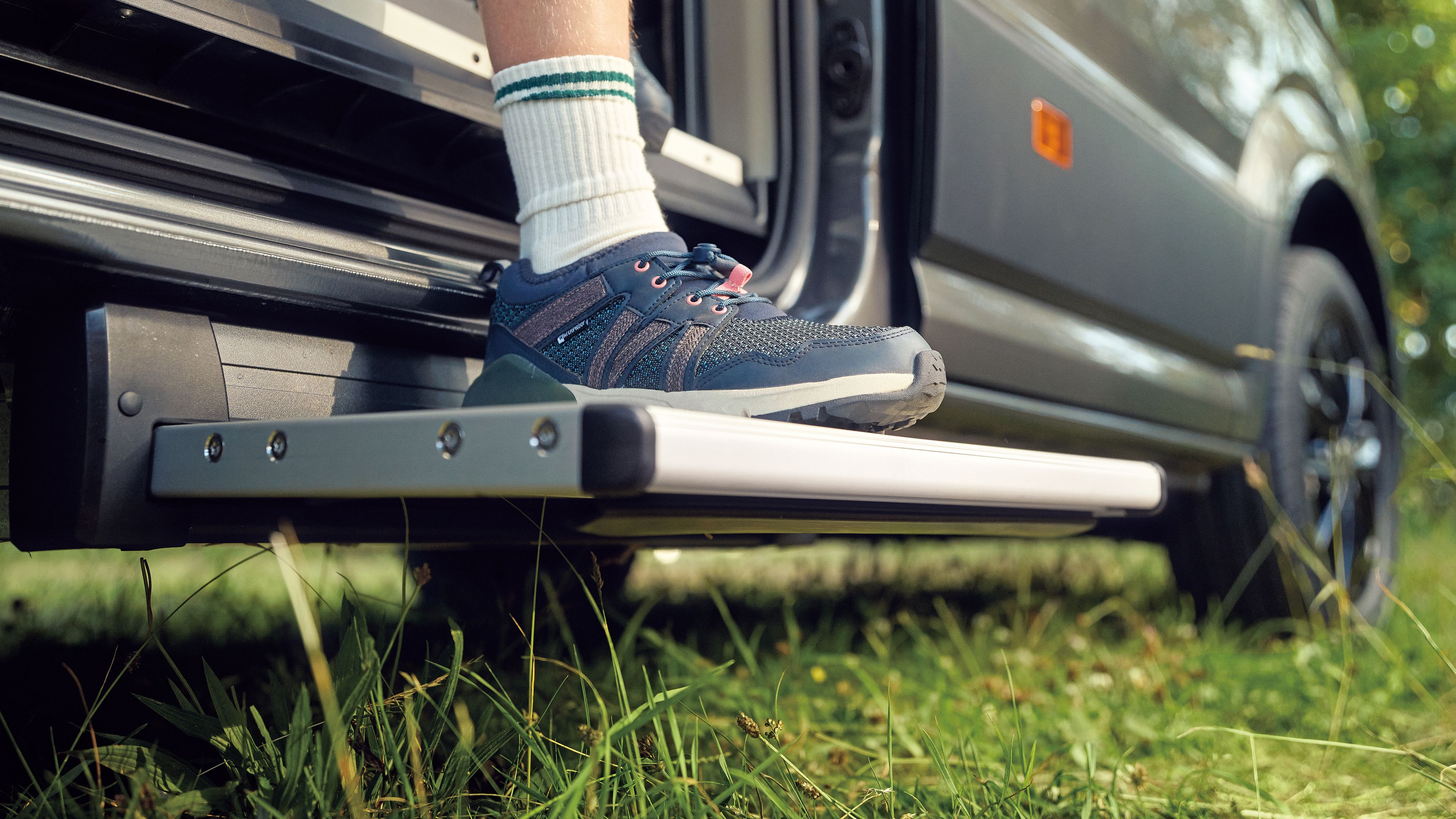 Eine Person mit weißen Socken und blauen Sneakern steht auf der ausziehbaren Trittstufe eines Campingwagens. Der Campingwagen steht in einer grünen Umgebung mit Gras. Die Trittstufe ist teilweise ausgefahren und ermöglicht einen bequemen Einstieg.