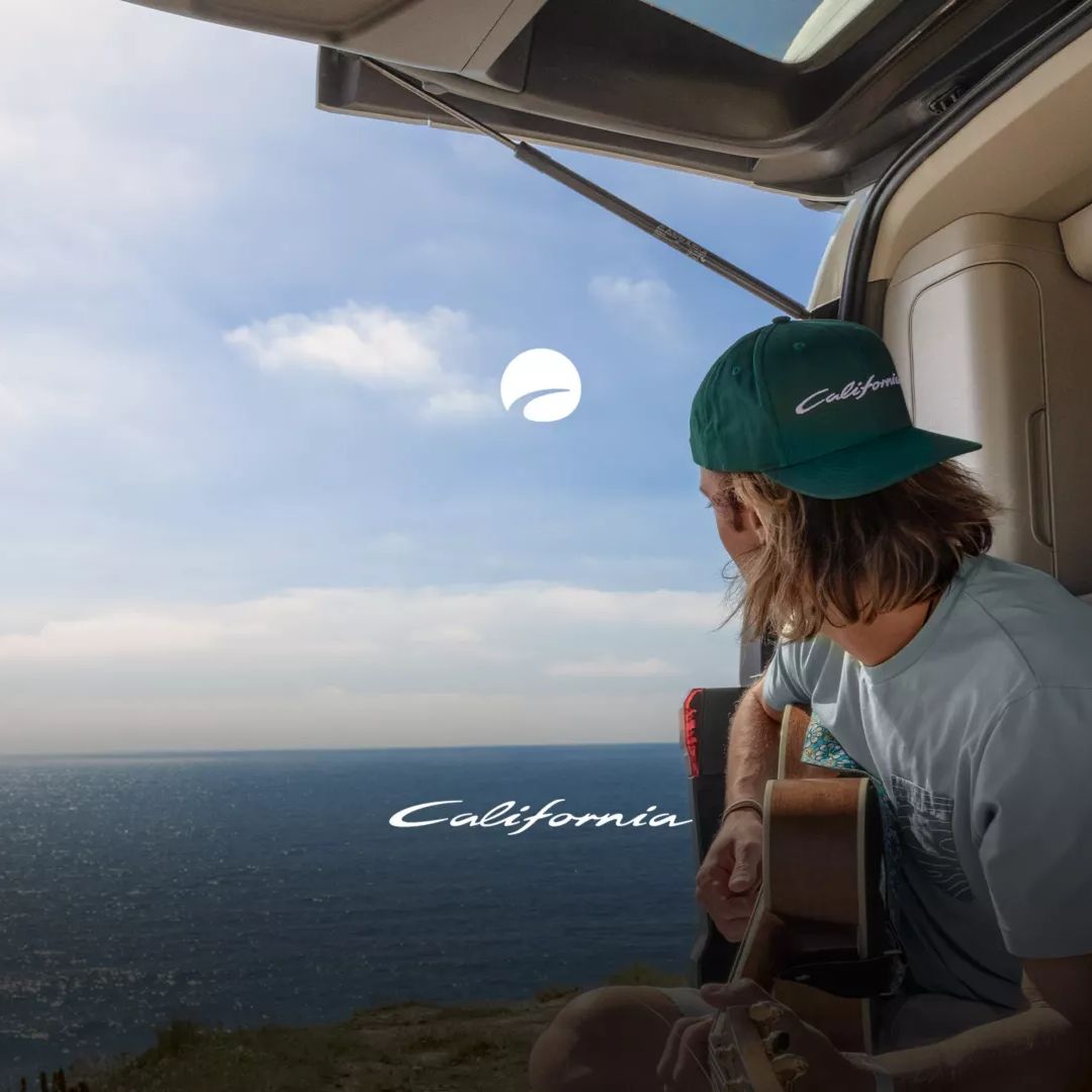 Ein Mann mit langen Haaren und grünem California Cap spielt Gitarre und blickt von einem Wohnmobil aus auf das weite Meer. Der Himmel ist blau mit wenigen Wolken. Unter ihm befindet sich der Schriftzug "California".