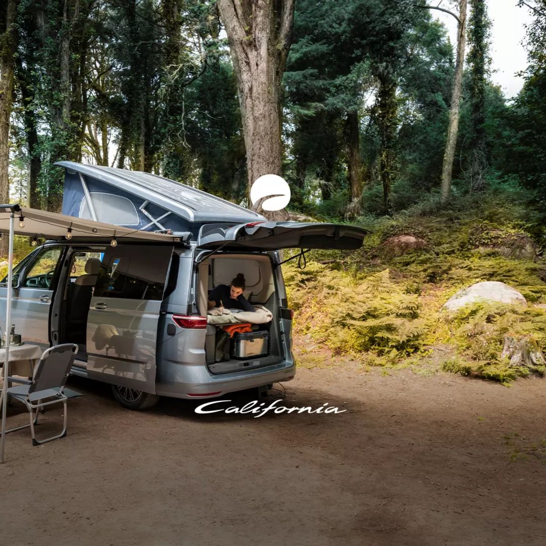 Ein VW California in einem Wald mit geöffnetem Aufstelldach, Campingtisch und Stühlen. Eine Frau liegt bei geöffnetem Heck im Fahrzeug. Auf dem Bild der California Schriftzug.