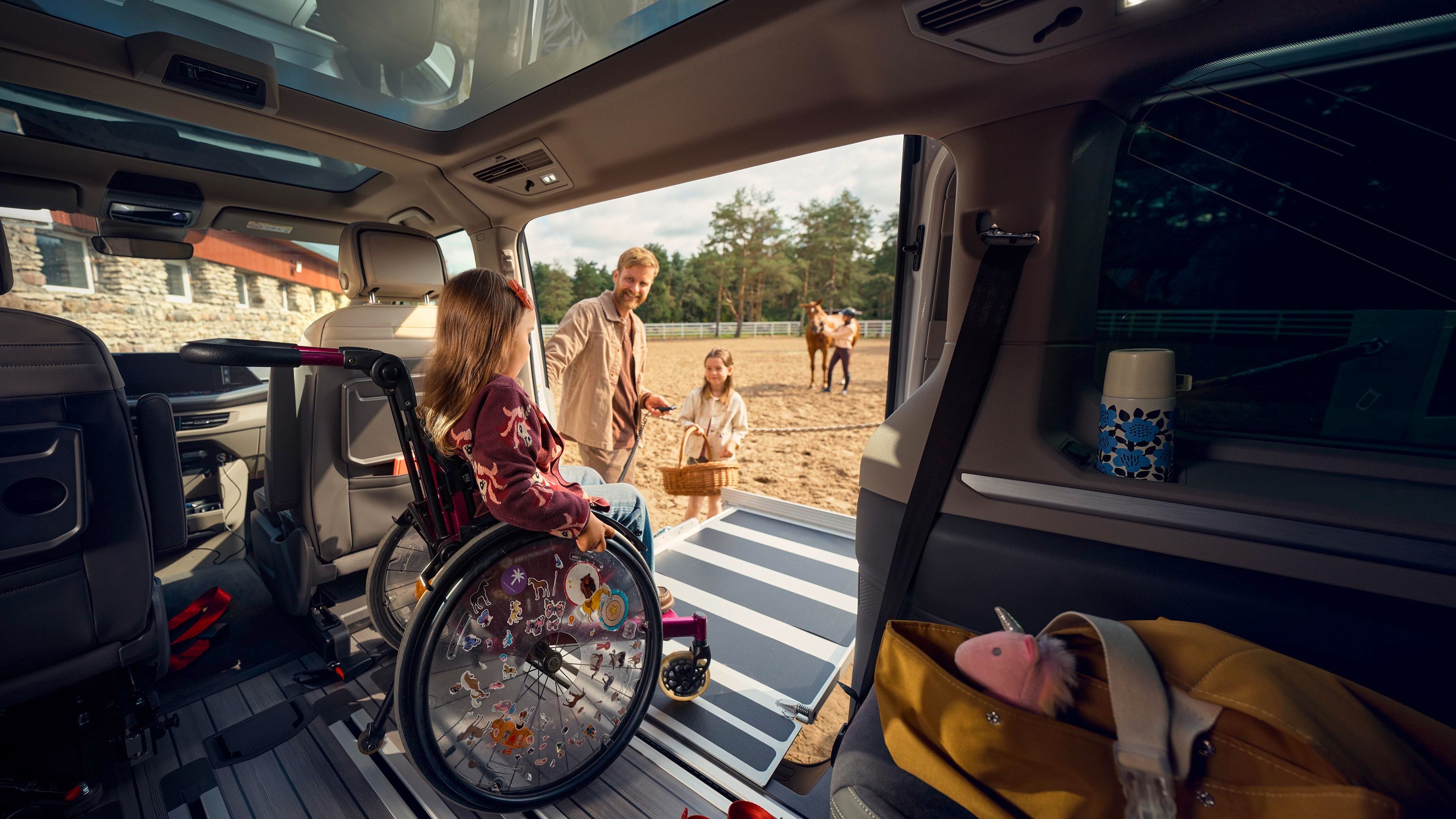 Ein Mädchen im Rollstuhl wird von einem Auto auf einen Sandplatz geschoben. Im Hintergrund sind zwei Erwachsene, die einen Korb tragen, sowie ein Pferd auf einer Koppel zu sehen. Die Innenseite des Autos ist teilmöbliert, einschließlich einer Tasche mit verschiedenen Gegenständen.