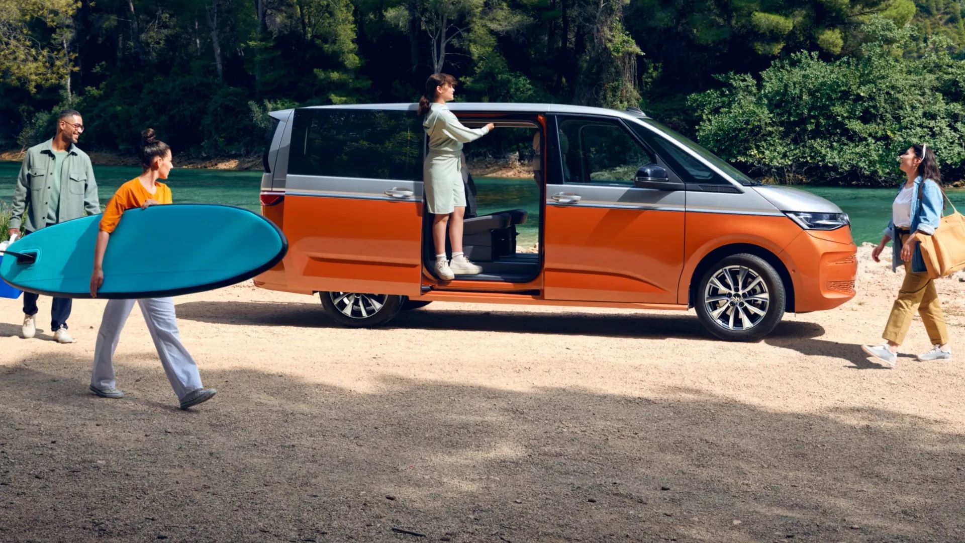 Seitenansicht eines VW Multivan mit geöffneter Schiebetüre an einem Fluss, eine Familie bewegt sich um das Auto