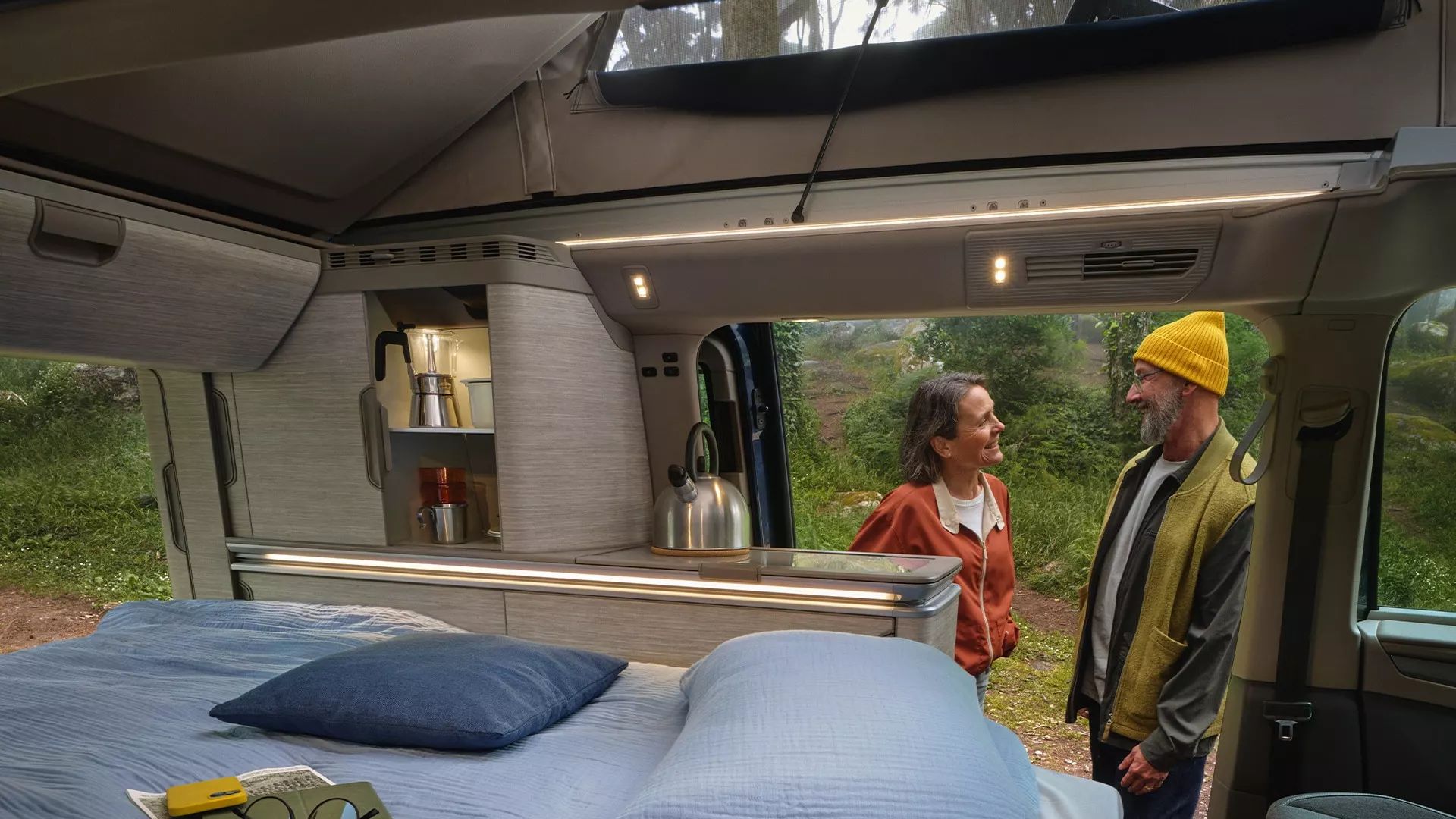 Der neue California Camper mit bezogenem Bett, gemütlicher Belichtung und 2 Personen unterhalten sich