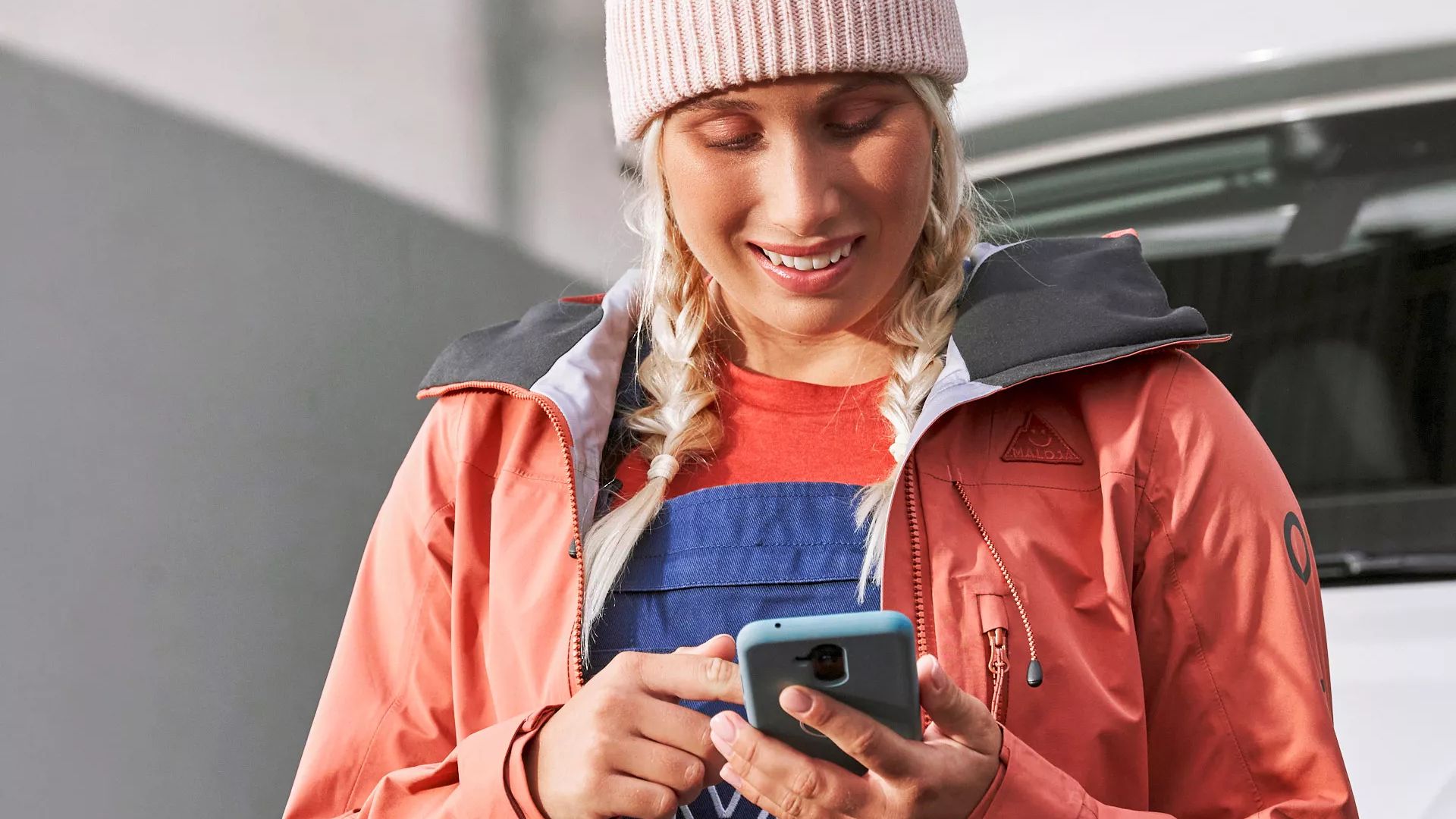 Eine Frau mit blonden Zöpfen und Mütze steht vor einem Wohnmobil und benutzt ein Smartphone. Sie trägt eine rote Jacke und blaue Arbeitshose. Ihr Gesicht ist verschwommen.