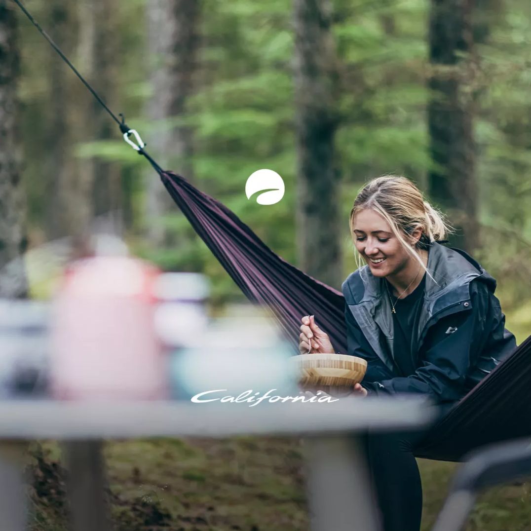 Eine Person sitzt in einer Hängematte und isst aus einer Schüssel, umgeben von Bäumen und einem geparkten Fahrzeug. Es sieht aus wie ein Campingplatz im Wald. Gegenstände stehen auf einem Tisch im Vordergrund.