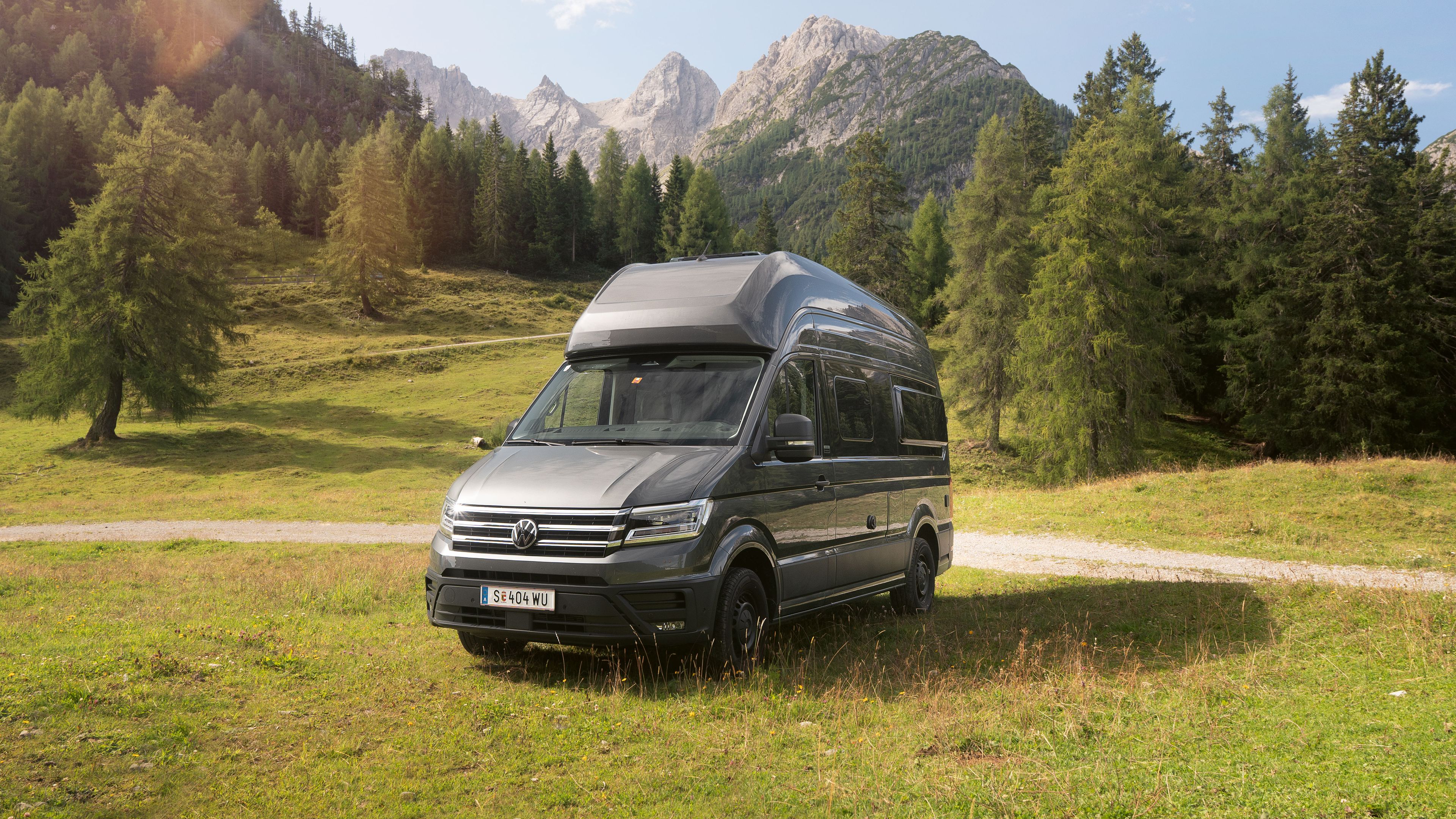 Ein silberner Wohnmobil-Van steht auf einer grünen Wiese vor einer malerischen Berglandschaft mit Bäumen und blauem Himmel.