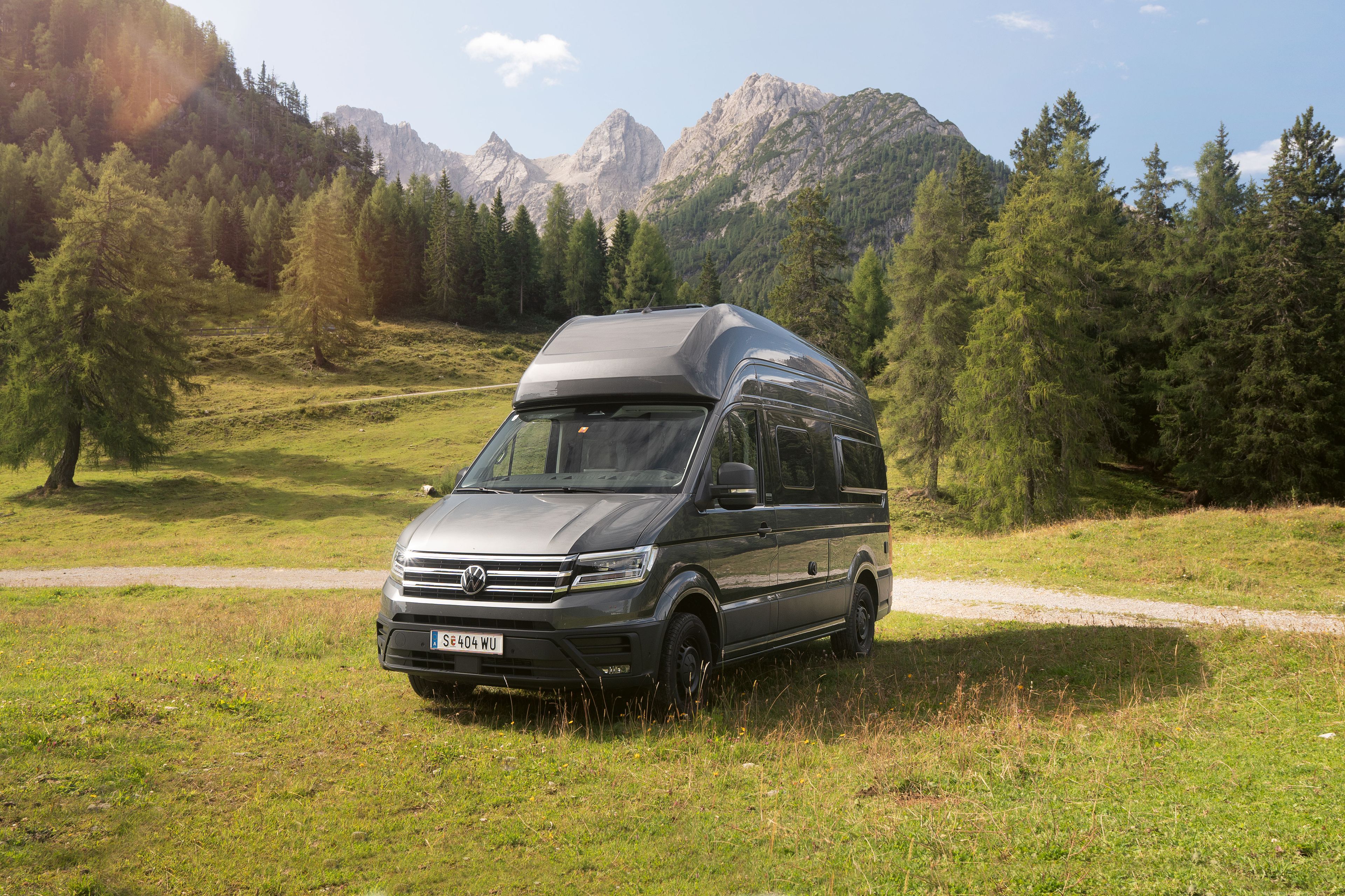 Ein silberner Wohnmobil-Van steht auf einer grünen Wiese vor einer malerischen Berglandschaft mit Bäumen und blauem Himmel.