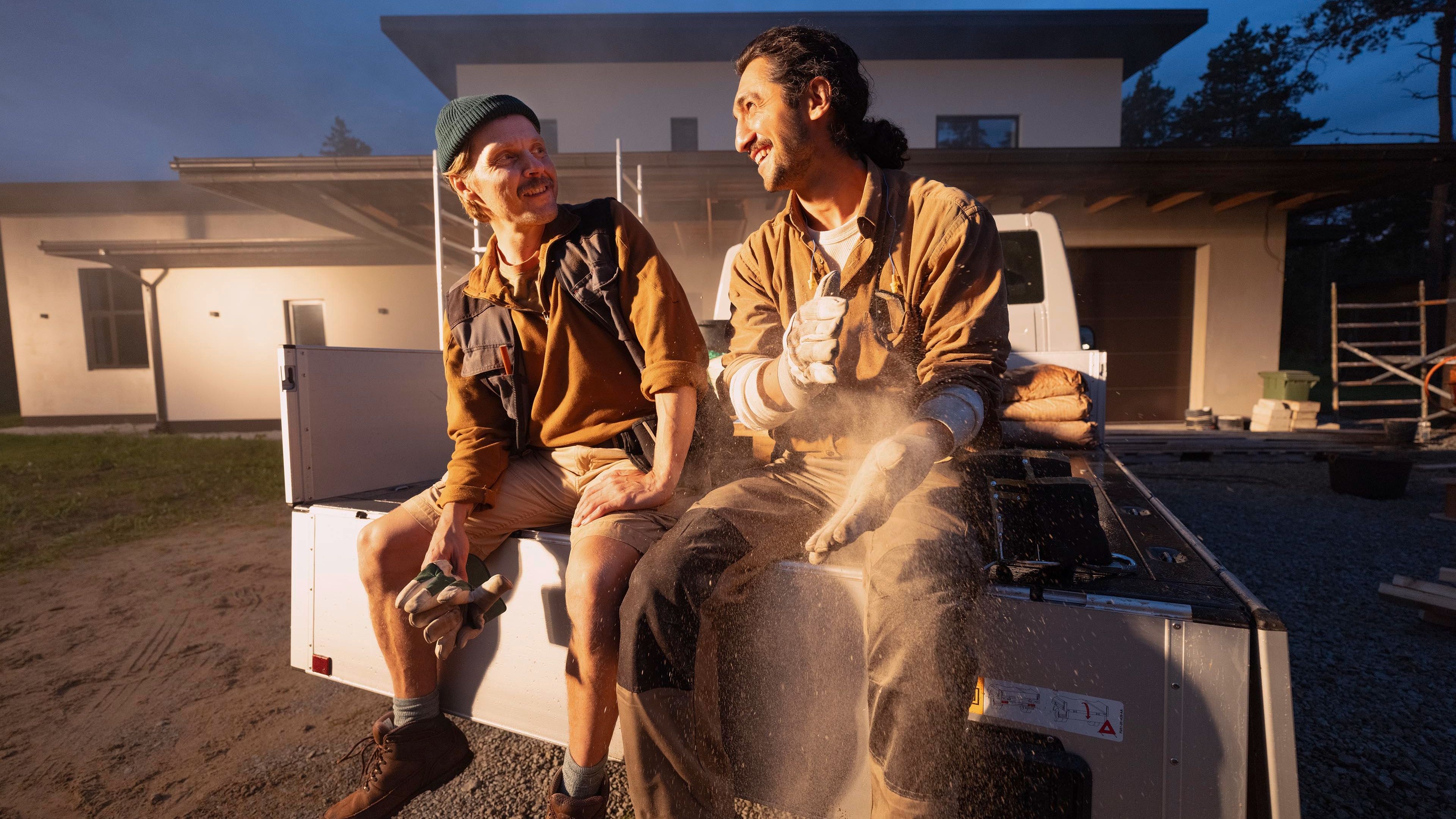Zwei Personen sitzen auf der Ladefläche eines Pick-up Trucks vor einem modernen Gebäude. Sie tragen Arbeitskleidung und schützen sich vor Staub. Die Umgebung scheint eine Baustelle zu sein.