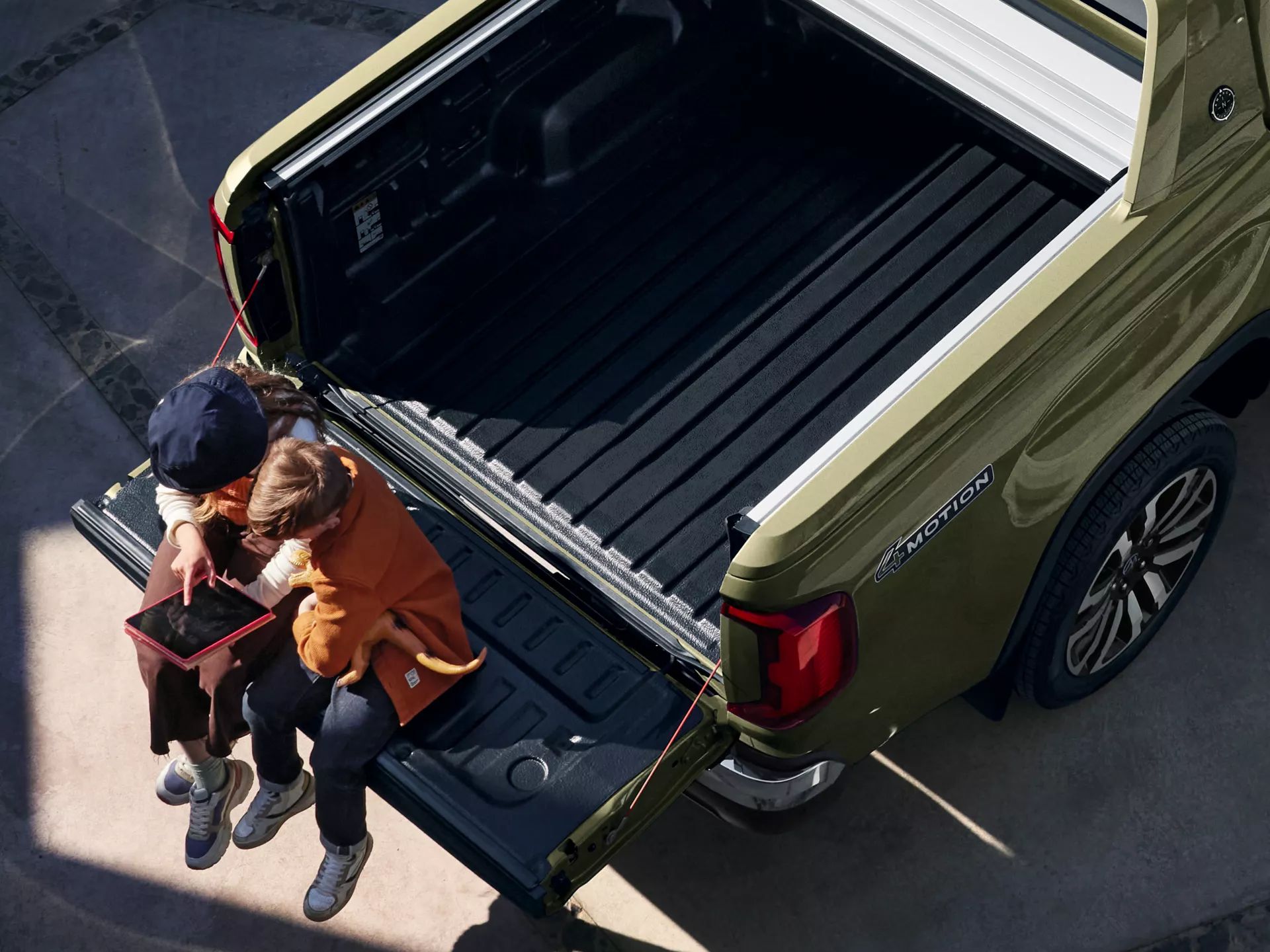 Zwei Kinder sitzen auf der heruntergeklappten Ladefläche eines olivgrünen Pickup-Trucks und benutzen ein Tablet. Die Ladefläche des Fahrzeugs ist leer und mit einer schwarzen Matte ausgekleidet. Die Szene findet draußen statt, bei natürlichem Licht.