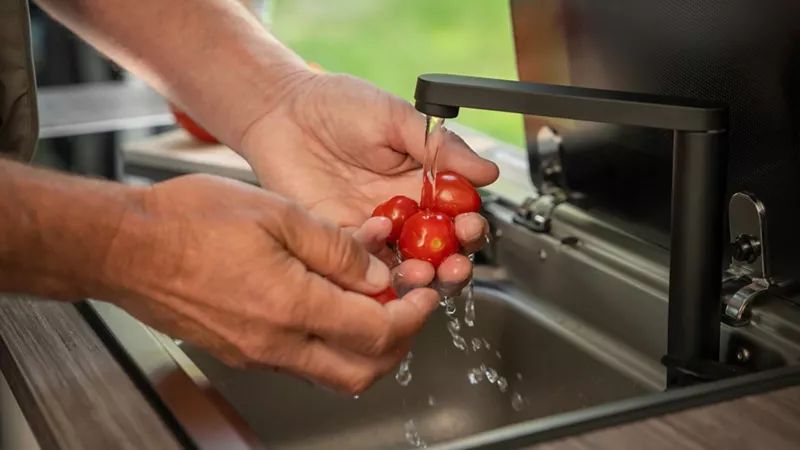 Zwei Hände halten mehrere kleine rote Tomaten unter fließendem Wasser in einer Küchenspüle. Die Tomaten werden gründlich gewaschen. Der Hintergrund zeigt verschwommenes grünes Laub.
