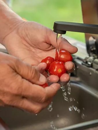Zwei Hände halten mehrere kleine rote Tomaten unter fließendem Wasser in einer Küchenspüle. Die Tomaten werden gründlich gewaschen. Der Hintergrund zeigt verschwommenes grünes Laub.