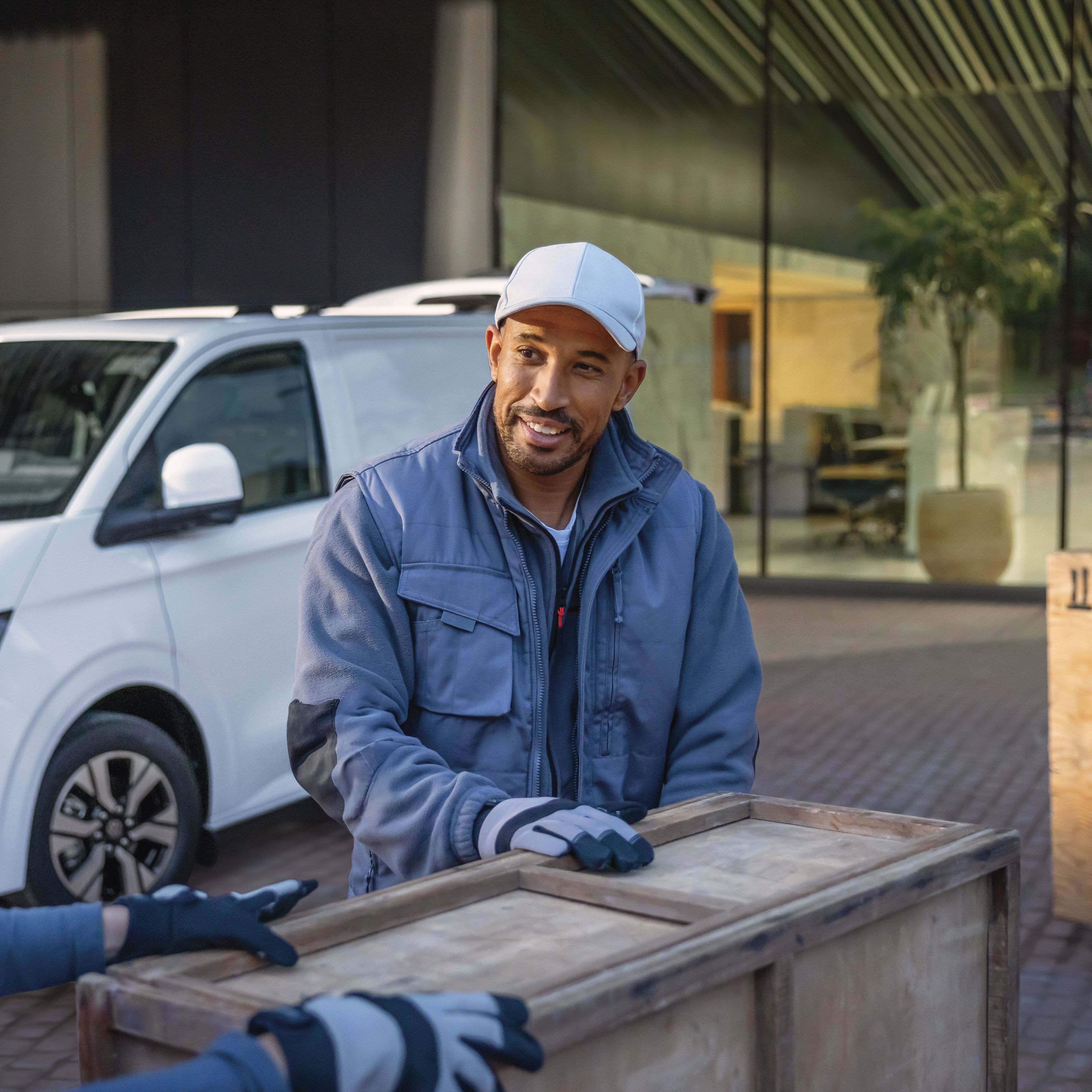 Zwei Männer tragen eine Holzkiste. Im Hintergrund ist ein weißer Volkswagen Transporter zu sehen.