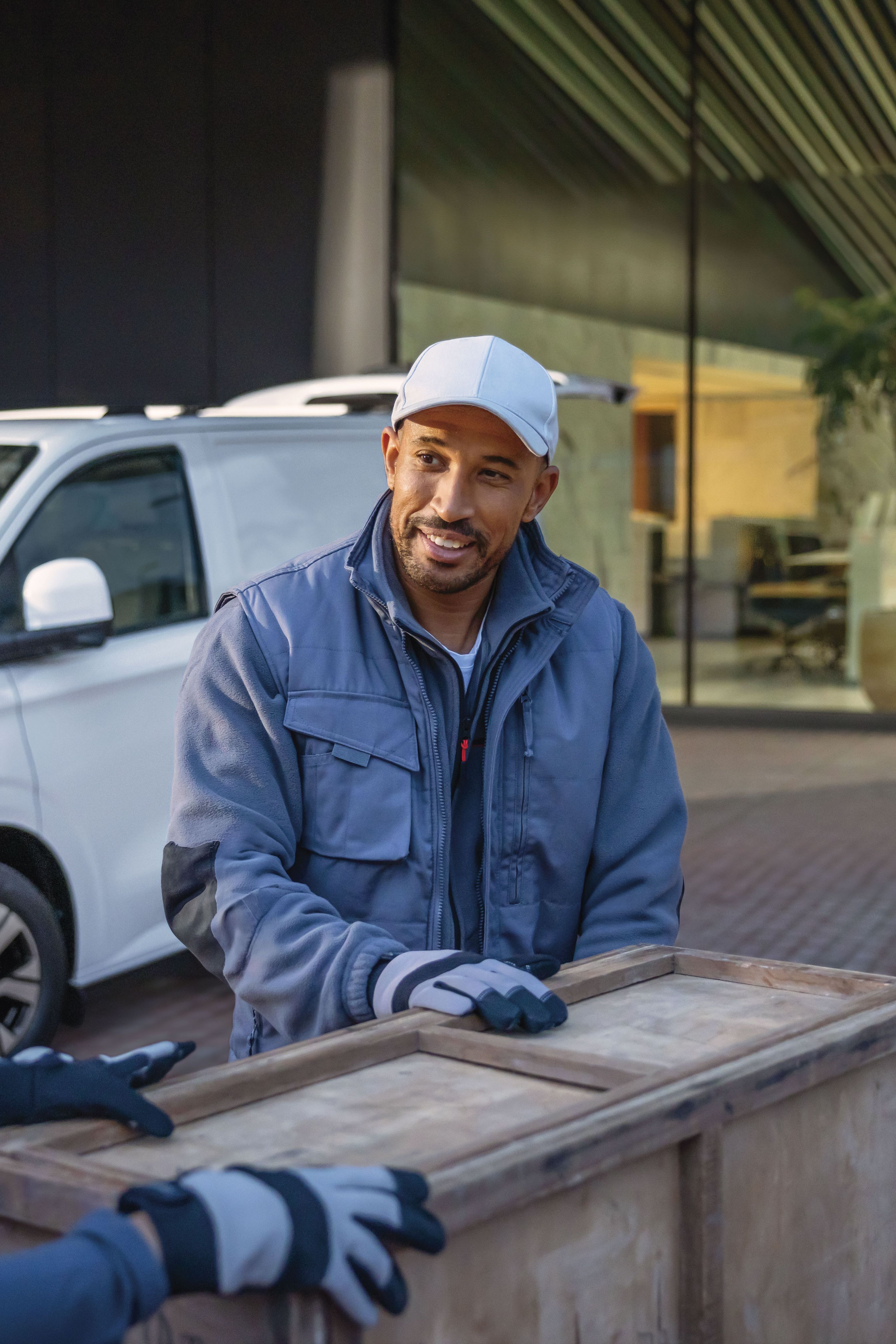 Zwei Männer tragen eine Holzkiste. Im Hintergrund ist ein weißer Volkswagen Transporter zu sehen.