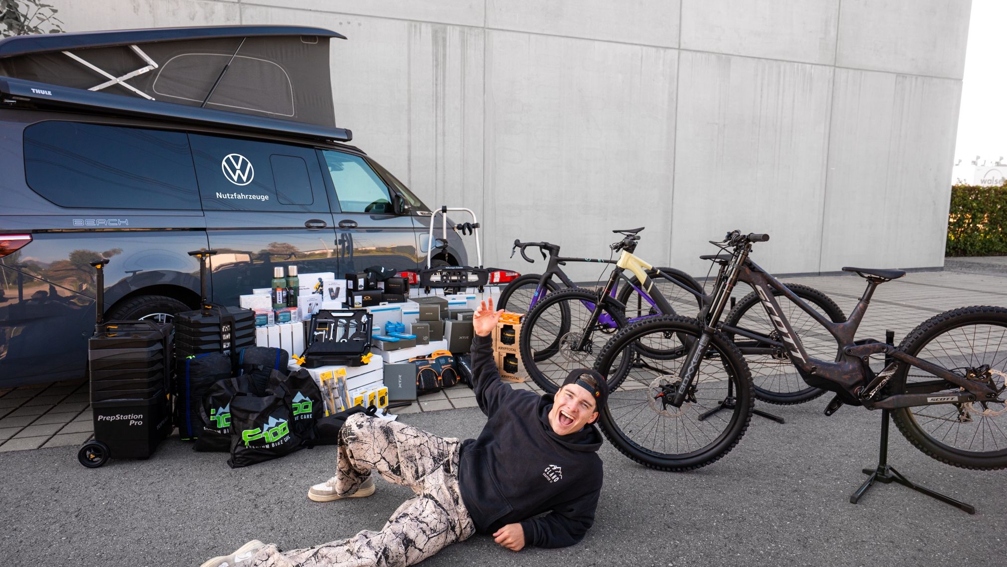 Ein Mann liegt vor einem Fahrzeug, das mit verschiedenen Fahrrad-Ausrüstungen beladen ist. Es sind mehrere Fahrräder sowie Zubehör wie Werkzeuge und Ersatzteile auf dem Boden ausgestellt. Der Mann scheint die Gegenstände zu präsentieren.
