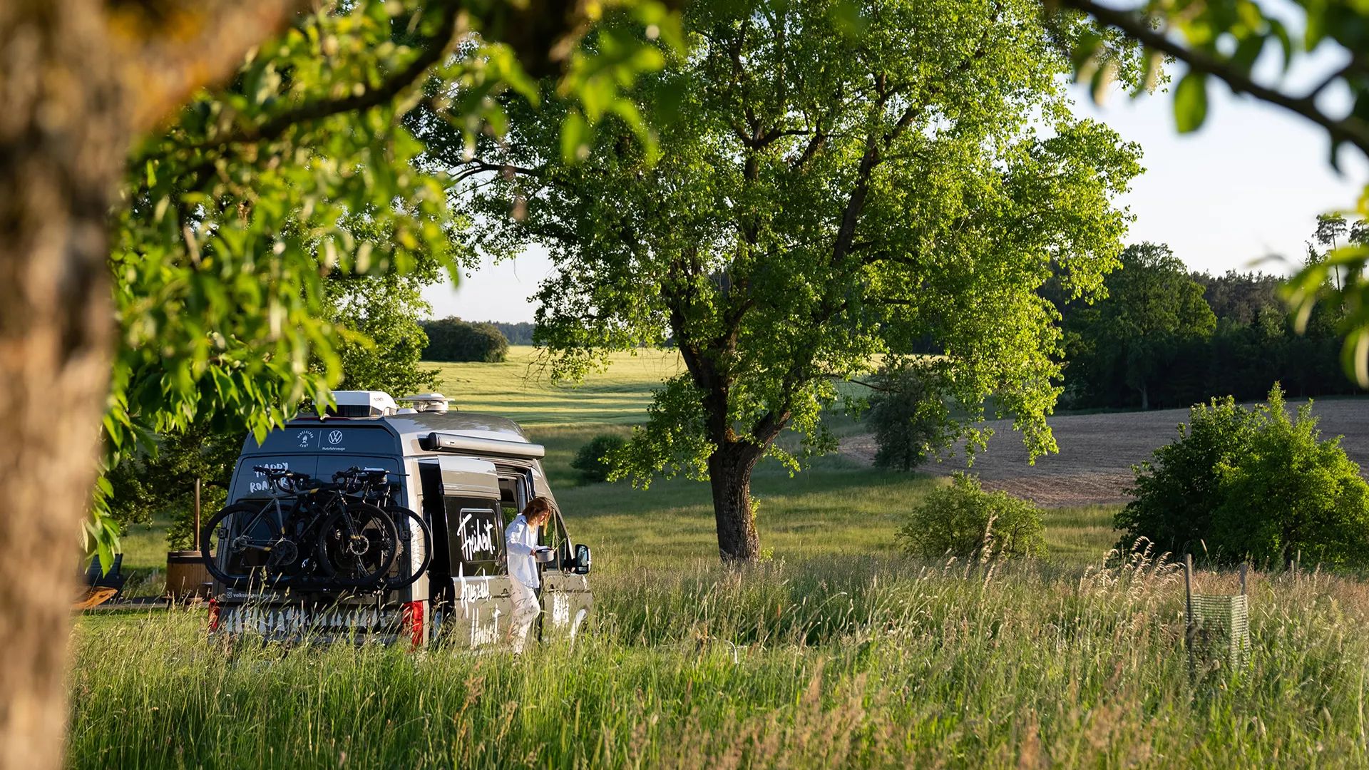 Ein Wohnmobil mit Fahrrädern am Heck steht auf einer grünen Wiese unter einem großen Baum. Das Wetter ist sonnig und die Landschaft im Hintergrund besteht aus weiteren Bäumen und Hügeln. Im Wohnmobil ist eine Person zu sehen.