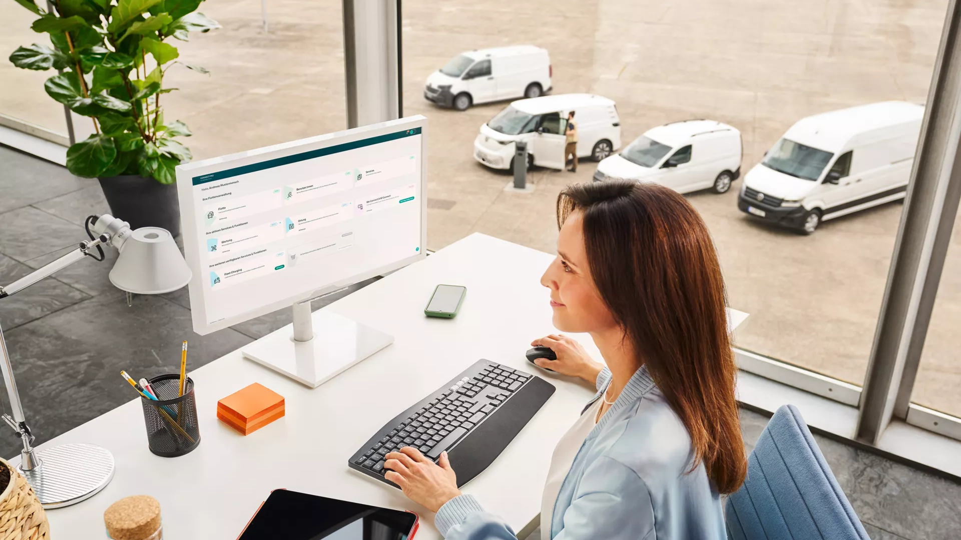 Eine Frau sitzt an einem Schreibtisch vor einem Computer in einem modernen Büro mit großen Fenstern. Draußen sind mehrere weiße Lieferwagen zu sehen. Auf dem Schreibtisch liegen eine Wasserflasche, ein Tablet und Schreibutensilien.