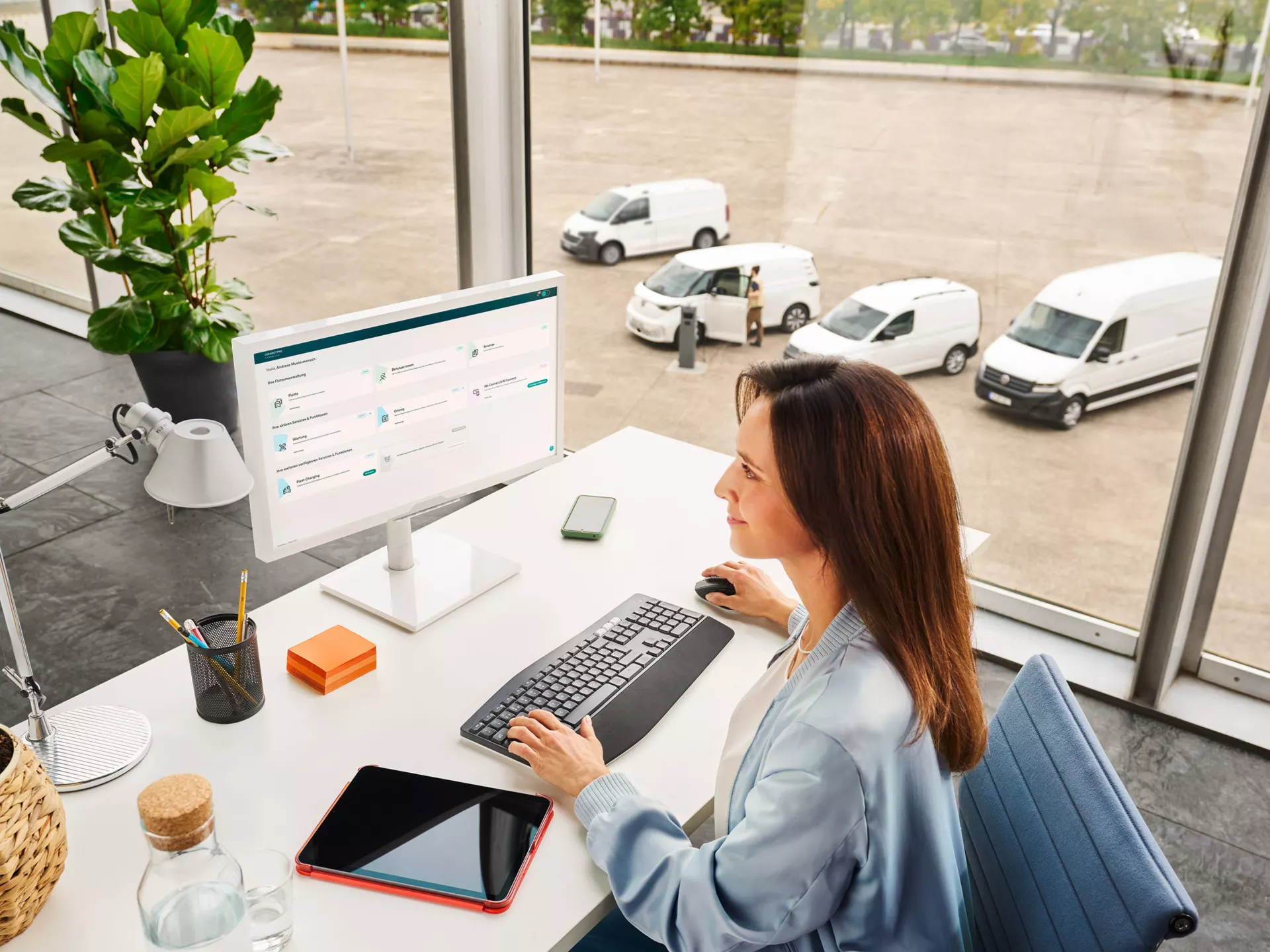 Eine Frau sitzt an einem Schreibtisch vor einem Computer in einem modernen Büro mit großen Fenstern. Draußen sind mehrere weiße Lieferwagen zu sehen. Auf dem Schreibtisch liegen eine Wasserflasche, ein Tablet und Schreibutensilien.