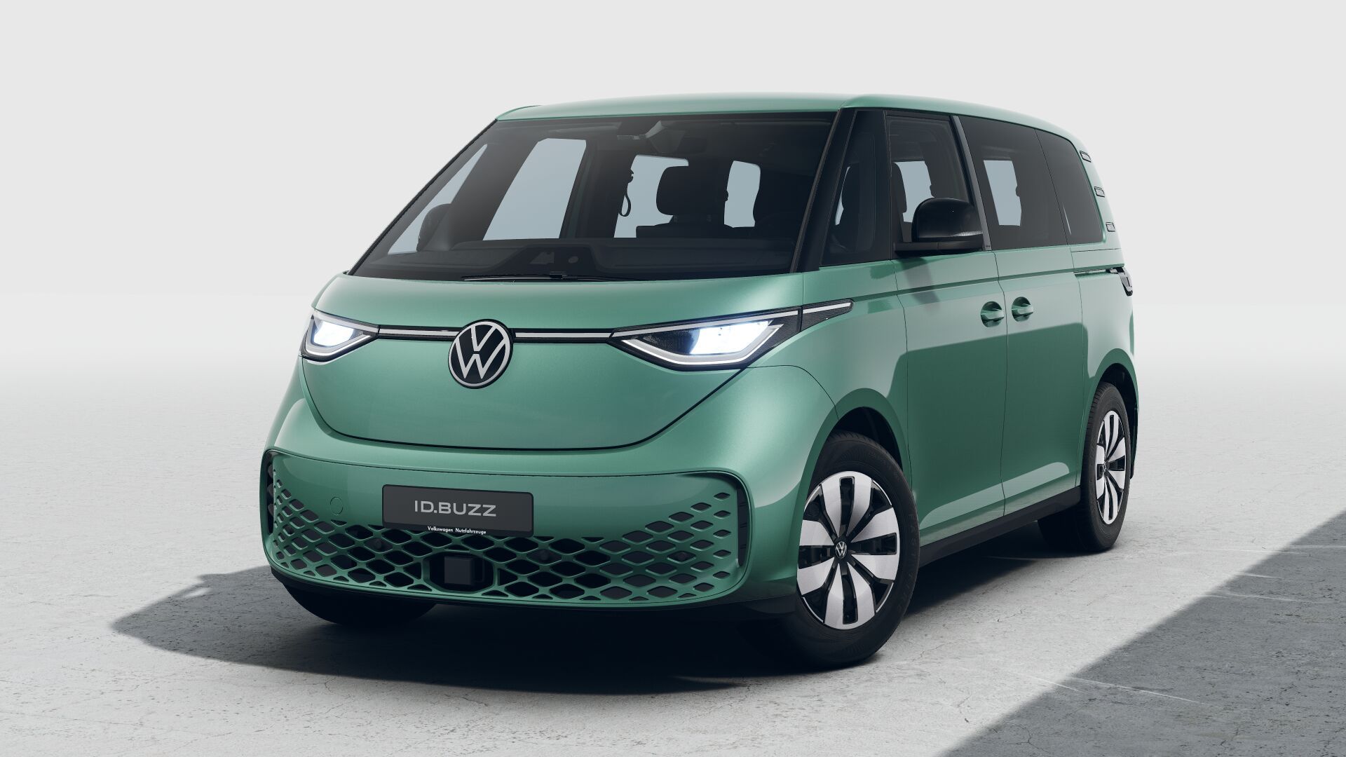 Grüner VW ID. Buzz, ein moderner elektrischer Kleinbus, steht leicht schräg von vorne fotografiert auf einer hellen Fläche. Die Front zeigt ein beleuchtetes VW-Logo, schmale LED-Scheinwerfer und eine auffällige, wabenförmige Struktur im unteren Stoßfänger. Die Seitenfenster sind groß und betonen den geräumigen Innenraumcharakter.