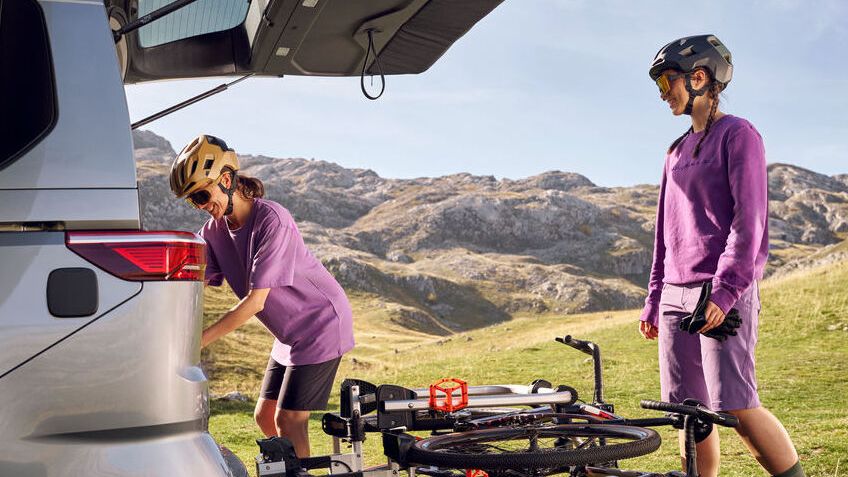 Zwei Radfahrerinnen in lila Sportkleidung vor einem geparkten Volkswagen Multivan mit geöffnetem Kofferraum in einer bergigen Wiesenlandschaft. Eine Person verstaut etwas im Kofferraum, während die andere daneben steht und einen Fahrradhelm trägt. Auf einem Heckträger ist ein zusammengeklapptes Fahrrad montiert.