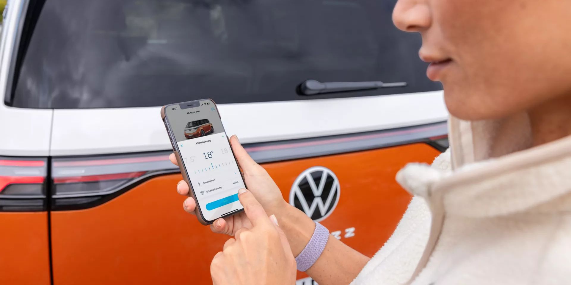 Eine Person benutzt eine Smartphone-App vor einem orange-weißen Volkswagen-Fahrzeug. Die App scheint Informationen oder Funktionen des Fahrzeugs anzuzeigen. Die Person trägt eine hellgraue Jacke und ein lila Armband.