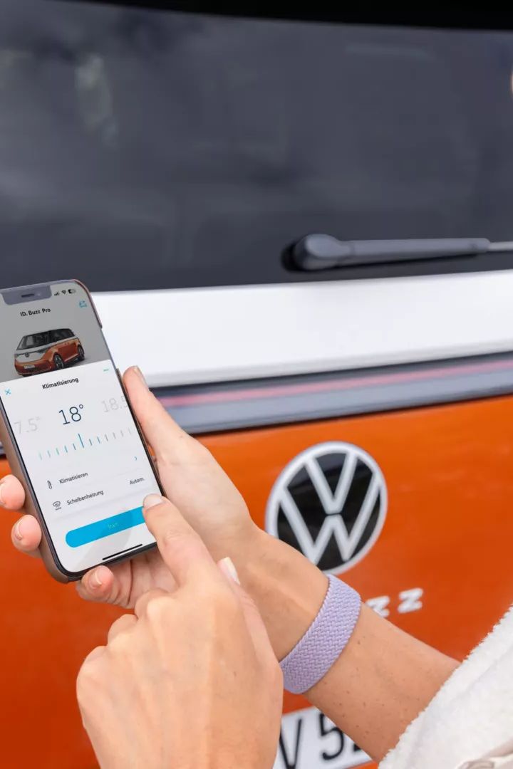 Eine Person benutzt eine Smartphone-App vor einem orange-weißen Volkswagen-Fahrzeug. Die App scheint Informationen oder Funktionen des Fahrzeugs anzuzeigen. Die Person trägt eine hellgraue Jacke und ein lila Armband.