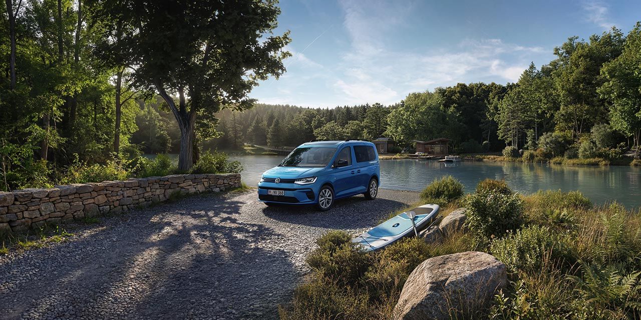 Ein blauer Volkswagen Caddy steht auf einem Schotterweg am Ufer eines ruhigen Sees, umgeben von dichtem, grünem Wald.