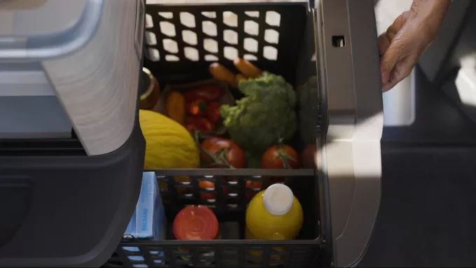 Ein Korb voller Lebensmittel inklusive Gemüse, Früchten und einer Saftflasche wird in ein Fach geschoben. Eine Hand hält die Fachklappe offen.