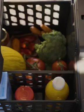 Ein Korb voller Lebensmittel inklusive Gemüse, Früchten und einer Saftflasche wird in ein Fach geschoben. Eine Hand hält die Fachklappe offen.